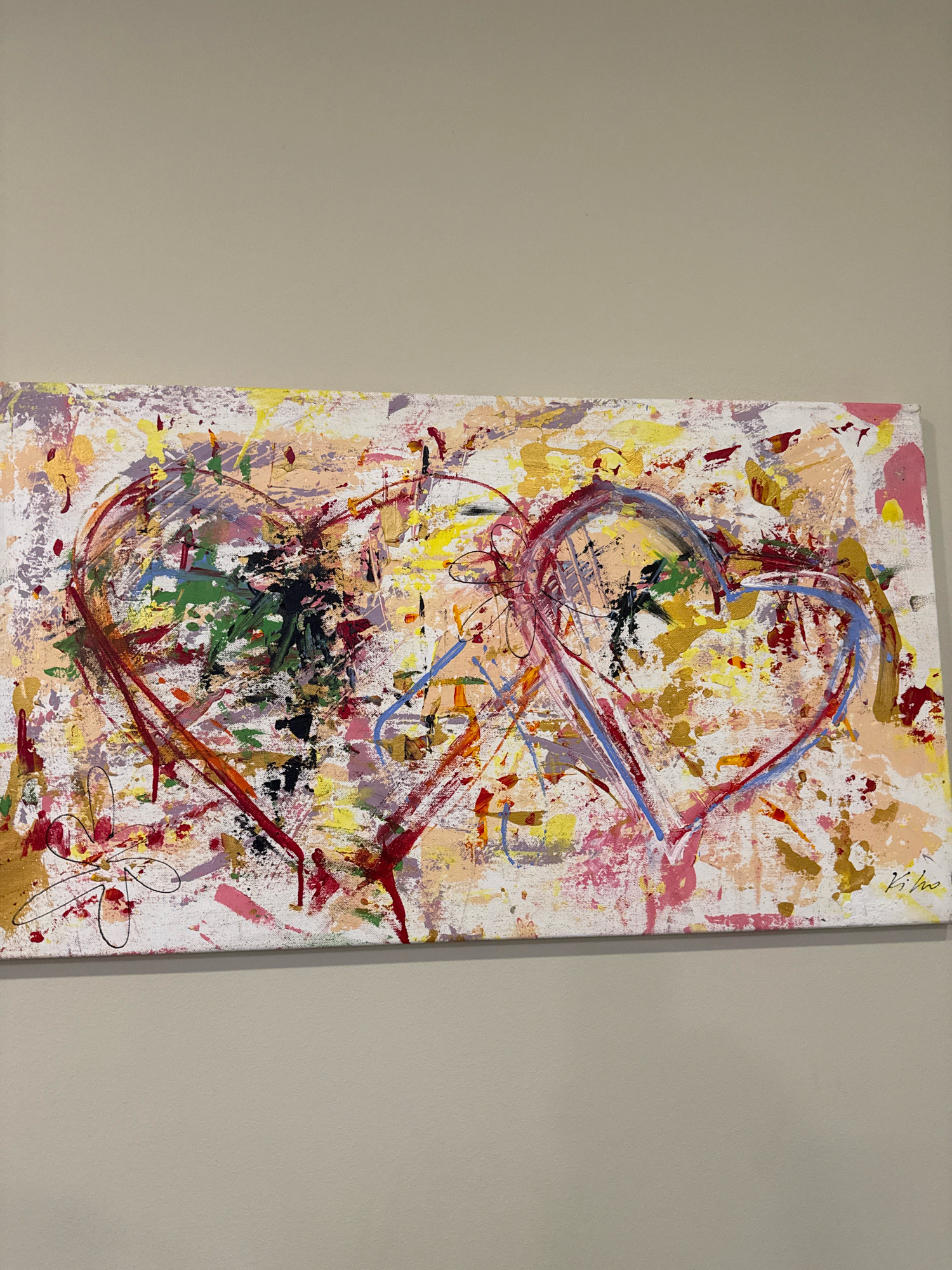 This is a photograph of an abstract painting of two intertwined hearts. The painting is rectangular and horizontally oriented, with a white background splattered with various colors including pink, yellow, orange, red, green, blue, purple, and black. Two large heart shapes are depicted in the center, outlined with bold brushstrokes in red and blue respectively, with some overlapping. The hearts are filled with energetic, abstract marks and textures. The painting is hanging on a neutral-colored wall, likely in a home or gallery setting, and is positioned centrally in the frame. The location in Tallinn, Estonia is not discernible from the image.