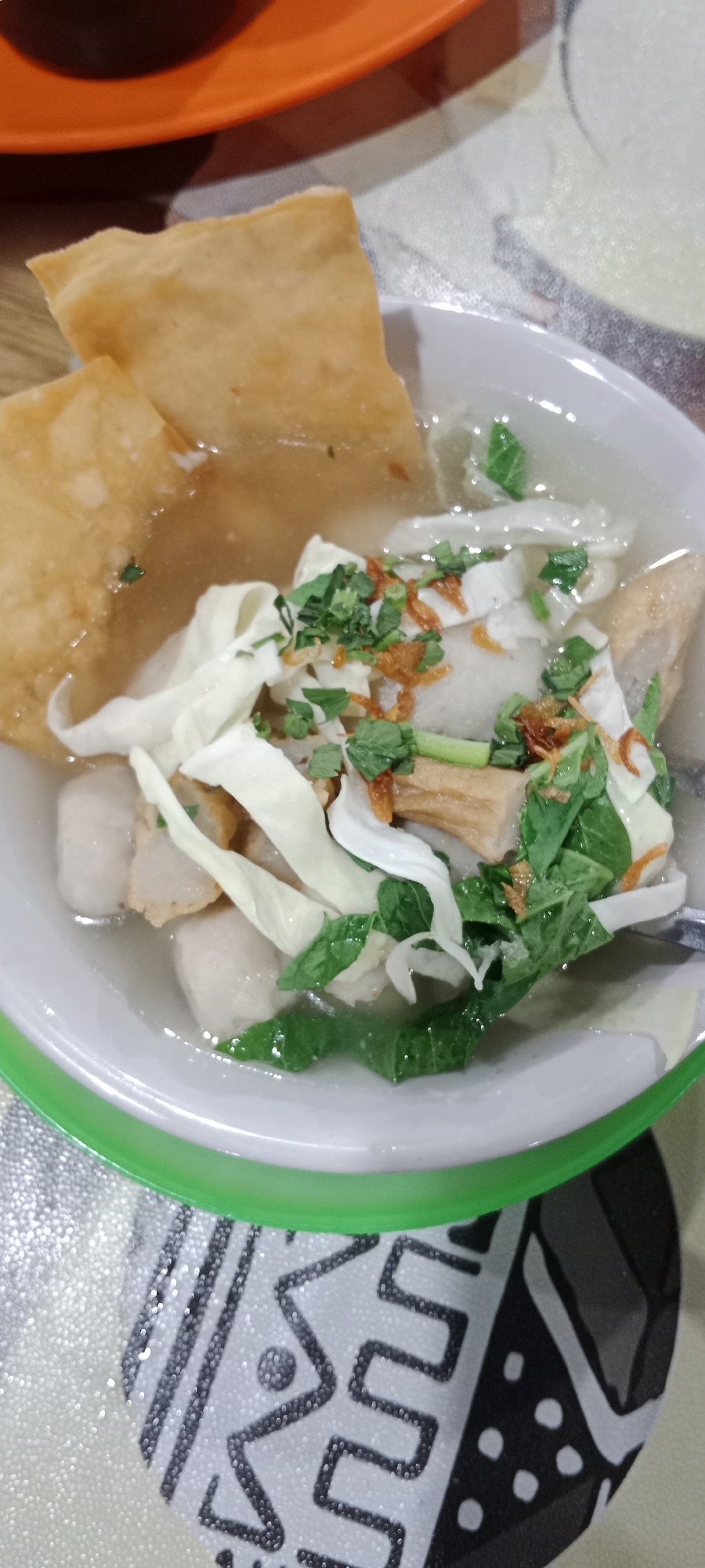 A close-up overhead shot shows a bowl of Indonesian food, likely bakso or a similar noodle soup, served in a white bowl with a green rim. The soup contains meatballs, sliced fried tofu or fish cake, shredded cabbage, and is garnished with chopped green herbs and fried shallots. Two pieces of golden-brown fried wonton or crackers are placed on top. The bowl sits on a patterned placemat with black and white geometric designs. An orange plate is visible in the upper background. This image was likely taken at a food stall or restaurant in Tanjung Pinang, Indonesia, a common setting for such dishes.