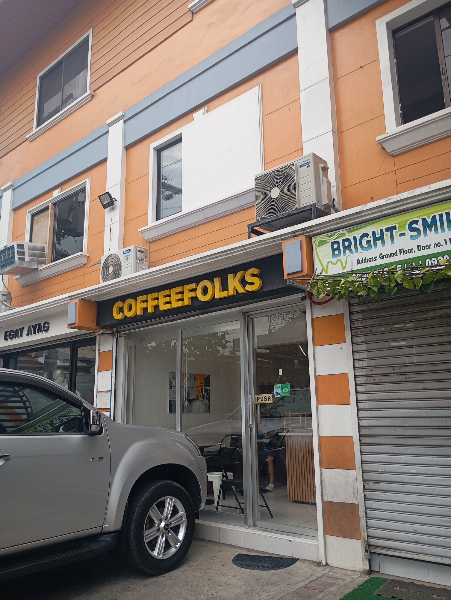 This image captures a street-level view of a commercial building in Davao City, Philippines, featuring multiple businesses. The building has an orange and white facade with grey accents, typical of urban architecture.
On the ground floor, a prominent yellow-on-black sign reads "COFFEEFOLKS," indicating a coffee shop with a glass entrance door. A "PUSH" sign and an "Open Grab Food" sticker are visible on the door. Inside, part of a person's leg is visible, suggesting someone is seated. To the left of COFFEEFOLKS, a partially visible sign says "EGAY AYAG." To the right, another business named "BRIGHT-SMILE" is indicated by a green and white sign with a tooth logo, likely a dental clinic. Underneath, the address "Ground Floor, Door no. 1" and a partial phone number "0920" are visible. This storefront is mostly covered by a metallic roll-down shutter, and artificial green foliage adorns its sign.
Several air conditioning units, including one branded "DAIKIN," are mounted on the building's exterior. The upper floors have multiple windows, some with screens, and parts of the building feature horizontally paneled siding.
A silver pickup truck, possibly an Isuzu D-Max given the visible "LS" trim badge on its side, is parked prominently in front of the coffee shop, with its front and side clearly visible. The truck has alloy wheels and appears clean.
The scene takes place during the daytime, characterized by bright, diffused lighting, possibly indicating an overcast sky. The overall impression is that of a bustling commercial area.