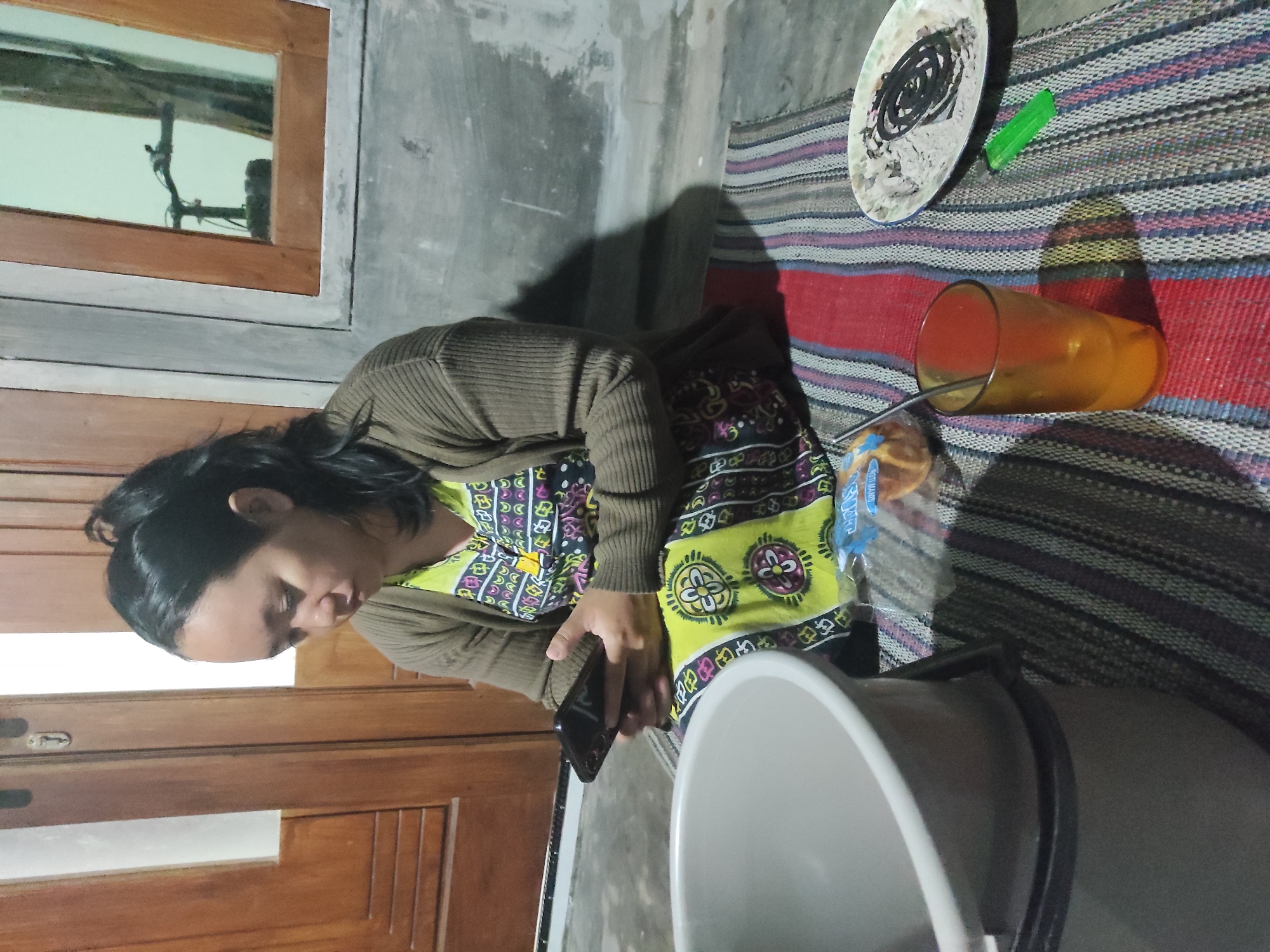 An Indonesian woman with dark hair pulled back in a ponytail sits on a colorful woven mat on the floor. She is wearing a brown cardigan over a brightly patterned yellow and black dress. The woman is looking down and holding a black smartphone in her hands. In front of her is a large gray plastic bucket, a glass with an orange liquid and a metal straw, a small plate with what appears to be a lit mosquito coil, and a small green object. Behind her, a wooden door with horizontal slats is visible, and to the right, a window with a wooden frame shows a reflection of a bicycle. The scene is likely in Kemuningsari Lor, Indonesia, based on the context of the prompt. The lighting suggests it is indoors and possibly evening.
