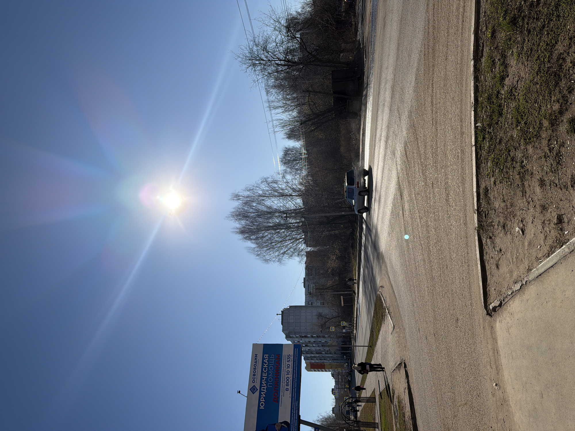 The image shows a street scene in Chaikovsky, Russia on a sunny day. The sun is bright, causing lens flare. A white vehicle is driving down the road, away from the viewer. On the left, a large billboard advertises "Юридическая помощь должникам" (Legal assistance to debtors) along with a phone number. Further down the road, buildings are visible, including an apartment complex. A person is walking on the sidewalk. The sidewalk is adjacent to a grassy area. The road surface is gray and appears slightly wet. Bare trees line the opposite side of the road. Power lines stretch across the top of the image.