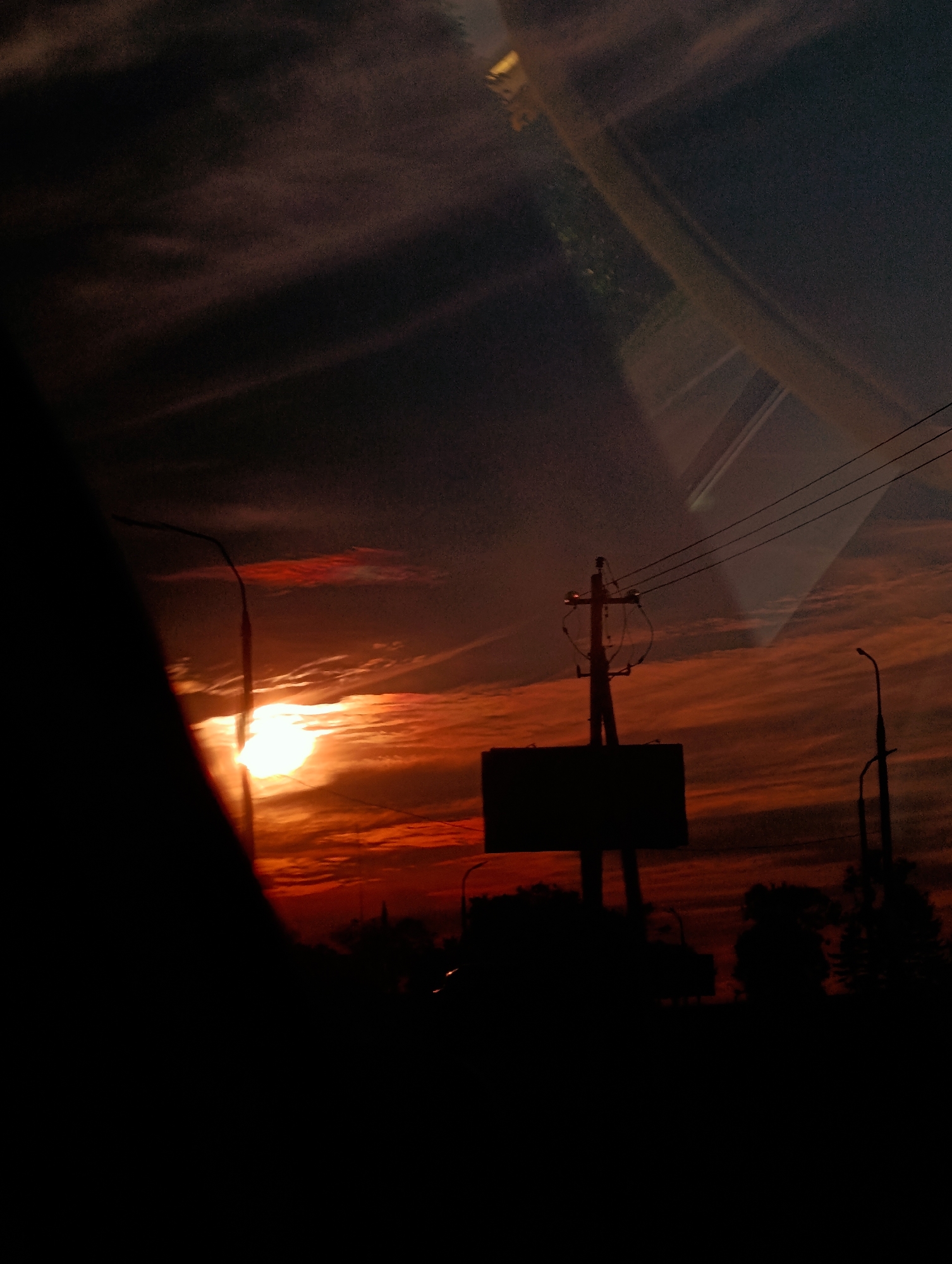 The image depicts a twilight scene, likely captured from a vehicle, with a striking sunset dominating the sky. The sun, a brilliant orb, is partially obscured by clouds and the silhouette of a street light pole in the foreground. The sky is a dramatic canvas of fiery oranges, reds, and deep purples, with wispy clouds creating a textured effect.
In the mid-ground, a dark rectangular sign is mounted on a pole, silhouetted against the vibrant sky. To the right, another street light pole is visible, also silhouetted. Power lines crisscross the upper portion of the image, contributing to the urban feel.
The foreground is dominated by the dark silhouette of what appears to be the interior of a vehicle, possibly a car window or dashboard, partially obstructing the view. This suggests the photograph was taken from inside a moving vehicle or from a stationary position looking out of a vehicle.
The lower portion of the image shows a dark, silhouetted landscape, likely trees and buildings, further emphasizing the transition from day to night. The overall impression is one of a dramatic sunset over a semi-urban or suburban landscape.
Based on the visual cues, such as the style of street lights and power poles, and the overall atmospheric conditions, this scene is consistent with a sunset in Minsk, Belarus. The specific location within Minsk cannot be determined from the image alone, but the elements suggest a road or roadside environment. There are no discernible people or specific actions taking place beyond the implied movement of the photographer or viewer.