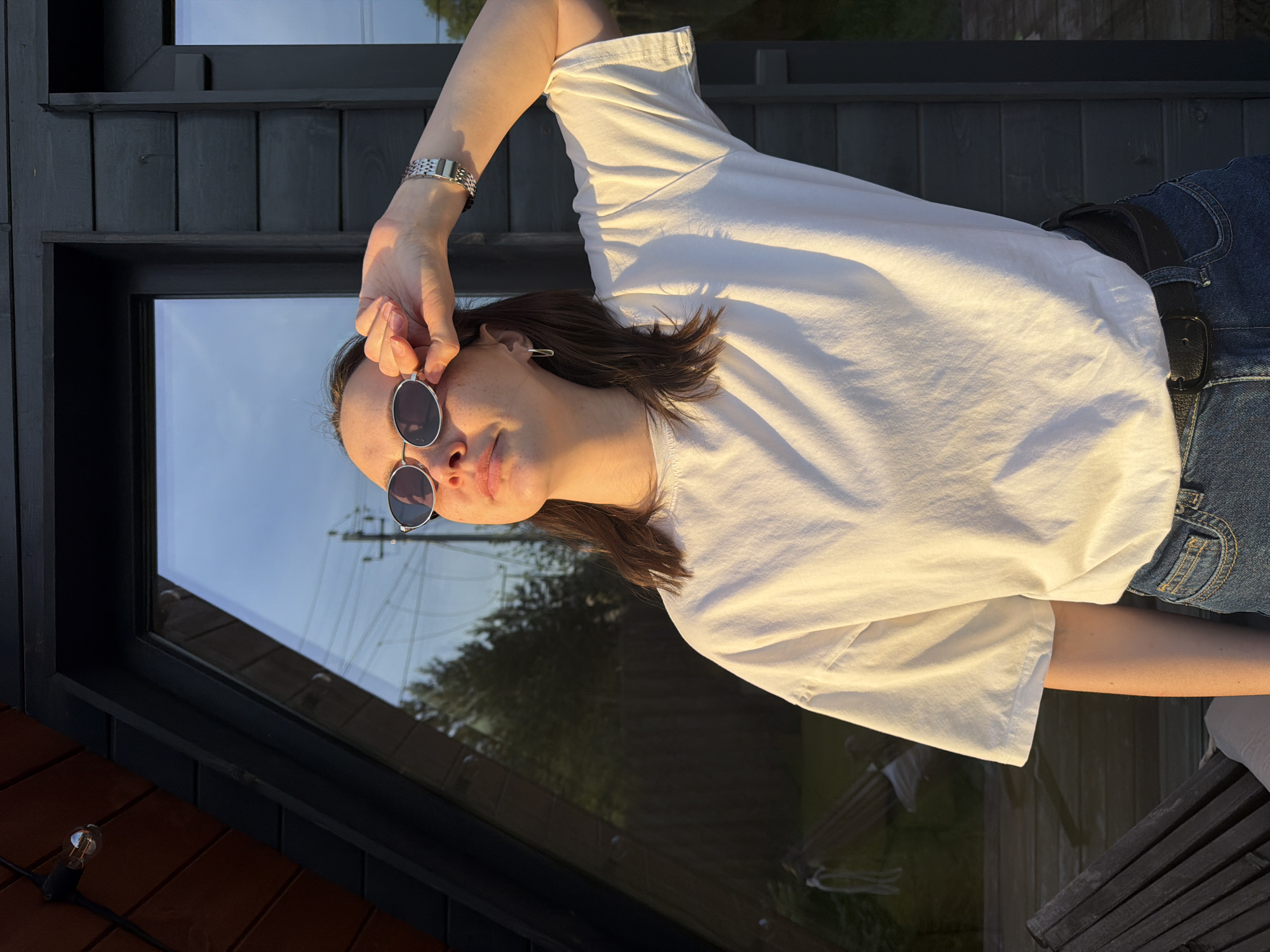 A woman is outdoors in what appears to be a backyard or deck area. She is wearing a white t-shirt, blue jeans with a black belt, and sunglasses. Her right hand is near her sunglasses. The background includes a building with dark wood siding and a large window, which reflects a view of the sky and trees. Outdoor string lights are visible near the corner of the building. The scene is bathed in warm sunlight. The exact location of Prikamskii, Russia is not discernable from the image.