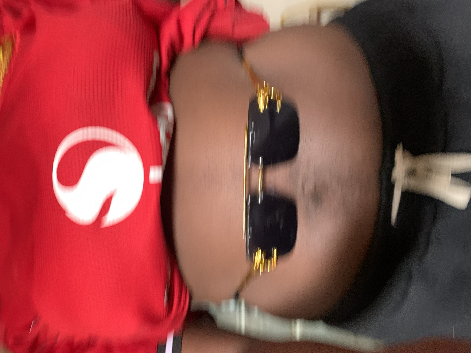The image displays a close-up, top-down perspective of a person, oriented vertically. The individual has dark skin and is wearing a vibrant red, ribbed shirt or jersey. A prominent white logo, appearing as a stylized 'S' or an incomplete swirl, is visible on the lower front of the shirt. The person is wearing gold-framed sunglasses with dark lenses, which obscure the eyes and upper face. Dark pants or shorts are visible on the lower body, with a partial view of a white strap or tie near the waist. The background is largely blurred, showing indistinct light brown and beige tones, suggesting an indoor or shaded setting. The person appears to be in a relaxed or reclining pose. No specific activity is depicted beyond posing for the photograph. Given the provided context, the image is situated at New Stadium, Nigeria, though no specific stadium elements are visible in the frame. No clear emotions, time of day, or weather details can be discerned.