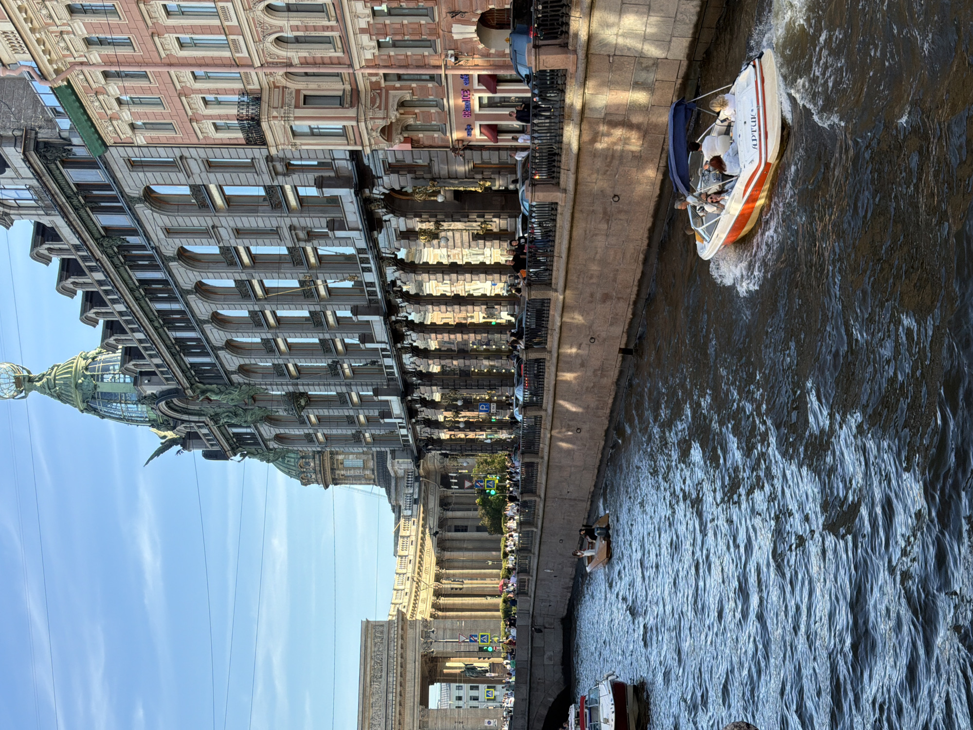 The image shows a view of the Griboyedov Canal in Saint Petersburg, Russia, looking towards the Singer House (Dom Knigi). Several boats are visible on the canal. In the foreground, a white and orange motorboat with a blue canopy is moving away from the viewer, carrying at least five people. Another smaller wooden boat with two occupants is further back on the canal. In the background, the Singer House with its distinctive green dome and globe is prominent. The canal is lined with ornate buildings with classical architectural features, including colonnades and arched windows. People are gathered along the embankment, some looking out at the canal. The sky is blue with some wispy clouds, and the sunlight casts shadows on the buildings and water, suggesting it is late afternoon or early evening.