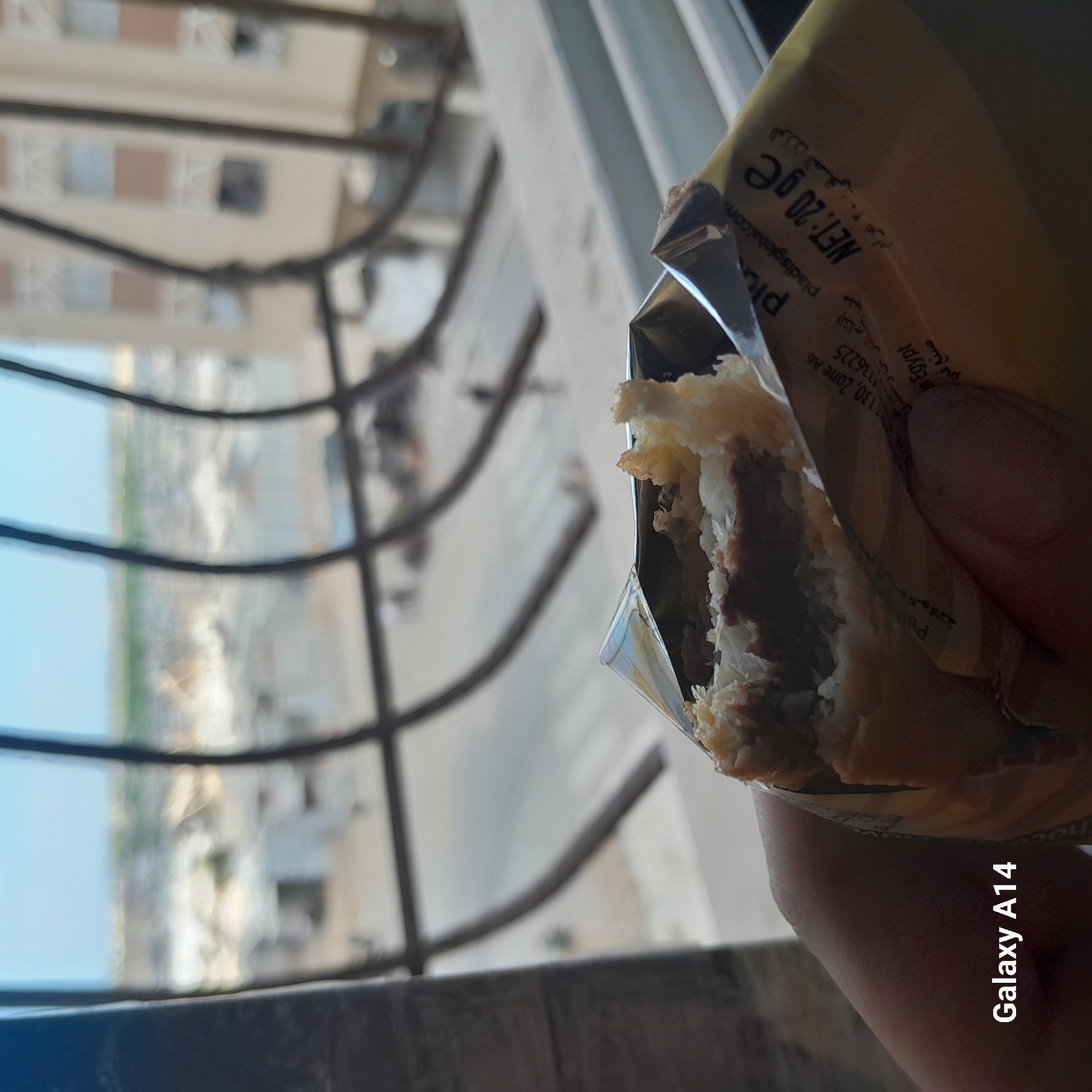 In Khan Yunis, Palestinian Territories, a close-up shot focuses on a partially eaten pastry held by a hand. The pastry, with visible layers of dough and a dark filling, is wrapped in a golden foil packaging. Text on the packaging, including "pladisglobal.com" and "NET: 20 ge," suggests it is a food item manufactured by Pladis. Arabic text is also present. Behind the pastry and hand, a blurred urban landscape is visible through a window with dark, curved bars. The landscape consists of buildings, possibly residential, and hints of open spaces. The sky above is a clear light blue. The overall impression is an ordinary scene of consumption against a backdrop of an urban environment.