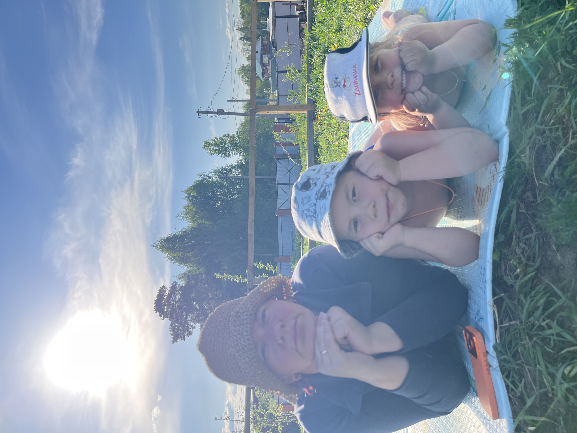 The image depicts three people lying on a light blue quilted blanket in a grassy area. The person closest to the camera is an adult wearing a brown crocheted hat and a dark-colored top. Their hands are positioned near their face. Above them is a child wearing a blue bucket hat, also resting their head on their hands. At the top of the image, another child wearing a white visor-style hat is making a face, pulling their cheeks out with their fingers. The sun is visible in the lower left corner of the frame.
In the background, there are trees, power lines, and buildings, suggesting a residential or suburban area. The photograph was taken outdoors during the day.
While specific information regarding Chaikovsky, Russia, is not directly discernible from the visual data, contextual elements like the style of housing and vegetation could potentially align with environments found in that region.