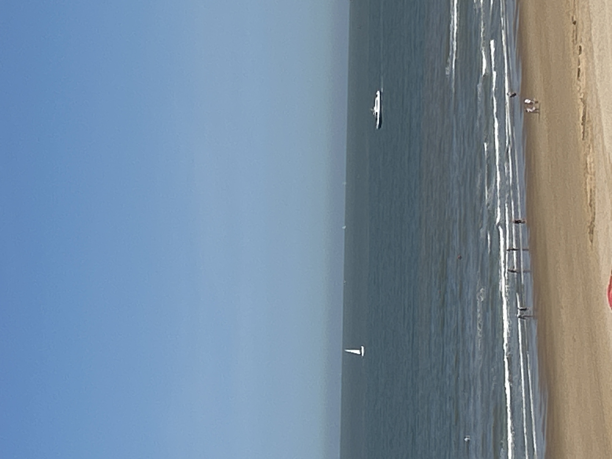 This is a photograph taken from a high vantage point overlooking the beach and North Sea in Ostend, Belgium. The sky is a clear, pale blue, with no clouds visible. The sea is a deep, muted blue, with gentle waves breaking on the shore.
In the mid-ground of the image, a lone white sailboat with its sail raised is visible on the water. Further to the right, a white and black motor yacht is cruising across the sea.
In the foreground, the sandy beach is sparsely populated. Several people are seen walking near the water's edge, with some appearing to be wading in the shallow waves. There is a hint of a red object, possibly a beach towel or umbrella, on the far right of the beach. The overall impression is a calm, sunny day at the coast.