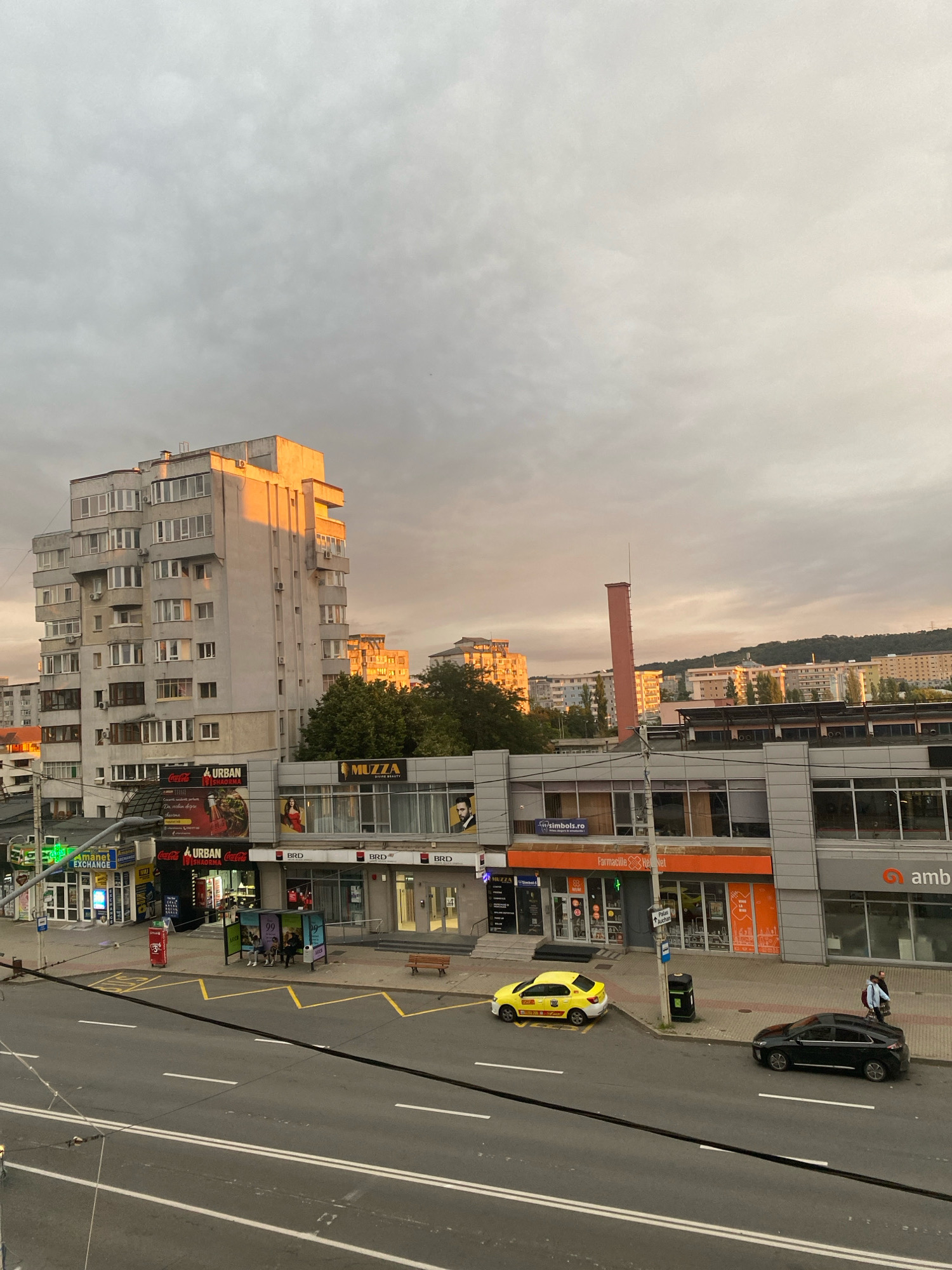 The image captures a street-level view of a commercial and residential area in Iași, Romania. The sky is overcast with hints of sunset light illuminating the upper parts of a tall, grey, multi-story apartment building on the left. This building features numerous windows and balconies, with some sections catching the warm glow of the sun. Below the apartment building, a row of commercial establishments lines the street. These include shops with prominent signage for "Coca-Cola," "URBAN," "MUZZA BEAUTY," and "BRD" (a bank).
Across the street, a yellow taxi and a black car are parked or driving. A bus stop shelter with people seated inside is visible on the sidewalk. The street itself is paved and marked with white lines, indicating multiple lanes of traffic. In the background, more buildings of varying heights and architectural styles are visible, some with orange-tinted facades, and a distant hillside covered with trees and structures. A tall, reddish-orange cylindrical structure, possibly a chimney or industrial element, stands out against the skyline. The overall scene suggests an urban environment during late afternoon or early evening.