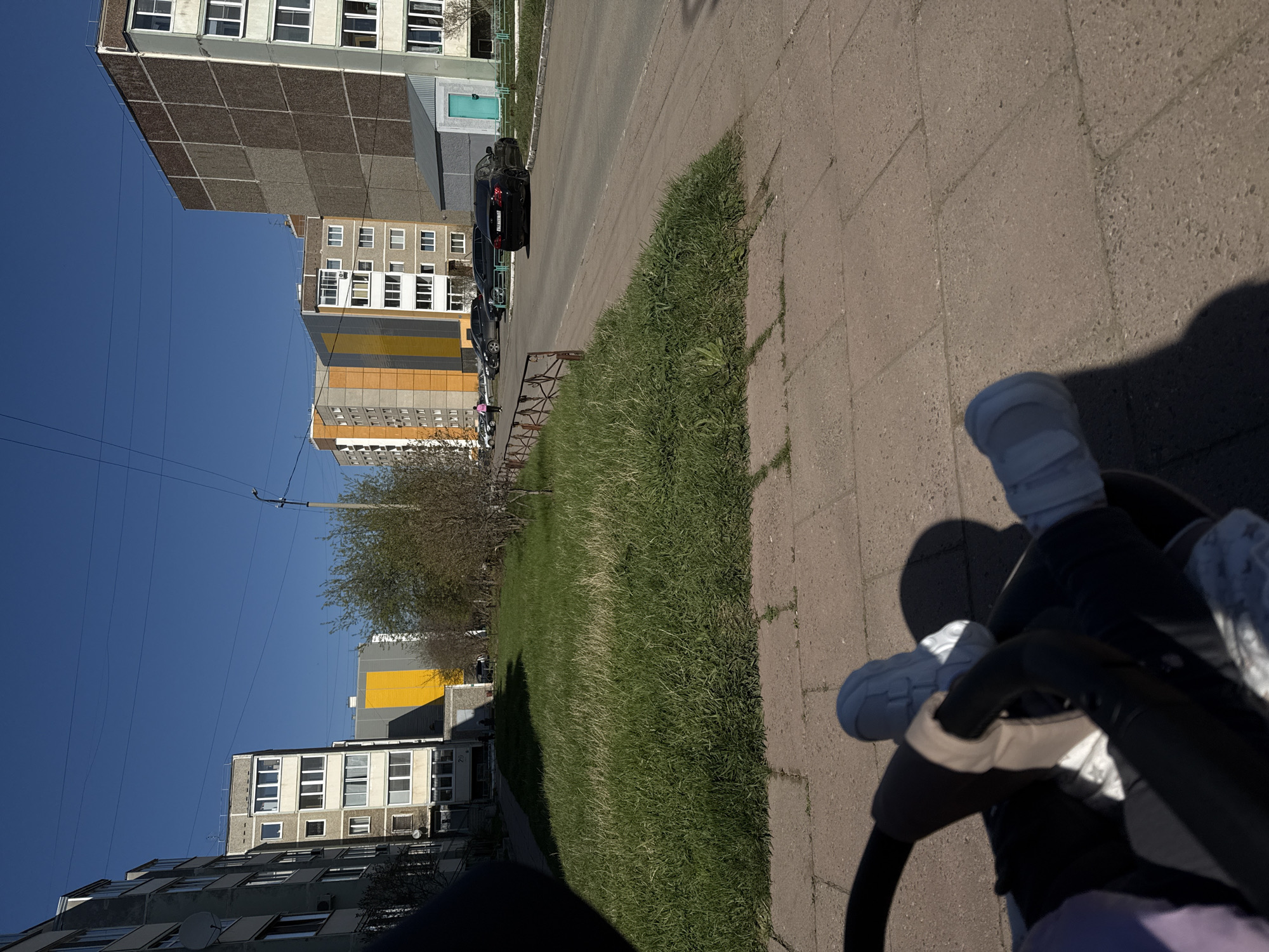 The image, taken in Chaikovsky, Russia, shows a view from what appears to be a stroller. A paved sidewalk runs along the right side of the frame. To the left of the sidewalk, there is a strip of green grass. Beyond the grass, several multi-story apartment buildings are visible against a clear, blue sky. Cars are parked along the street between the grass and the buildings. In the lower right corner of the frame, parts of the stroller are visible, including the handle covered with a white material.