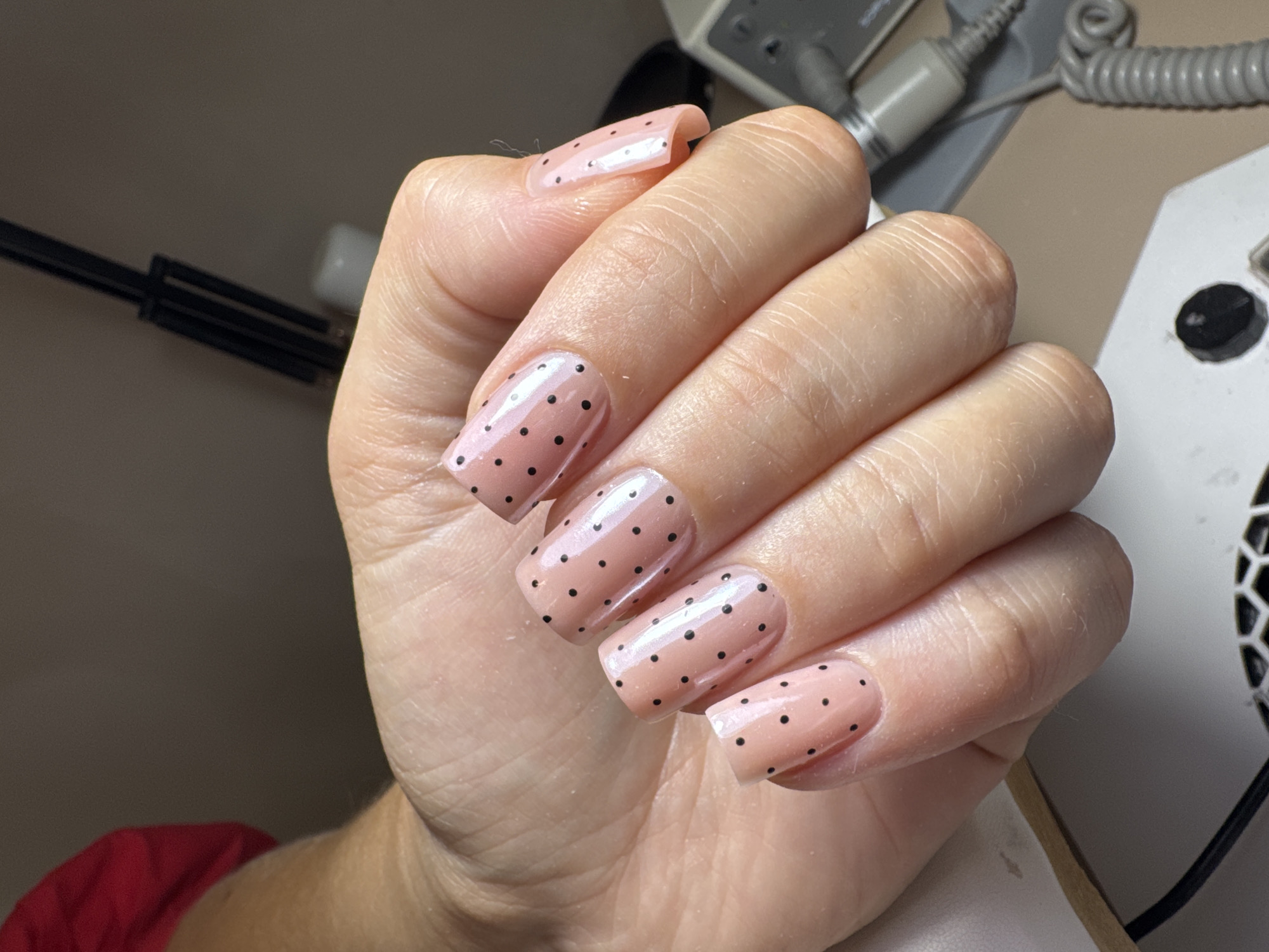 A hand with light pink square-shaped nails, each adorned with small black polka dots, is positioned against a cluttered background of manicure equipment and work surfaces. The skin on the hand is light-toned and shows creases and wrinkles consistent with age. The nails appear glossy.
The background includes a piece of red fabric in the lower left, possibly clothing. Manicure devices, like a drill with a coiled cord, are visible on a light-colored table or countertop. Other tools and a black flexible lamp are also present.
This specific location in Podgorica, Montenegro, cannot be determined from the given visual information.