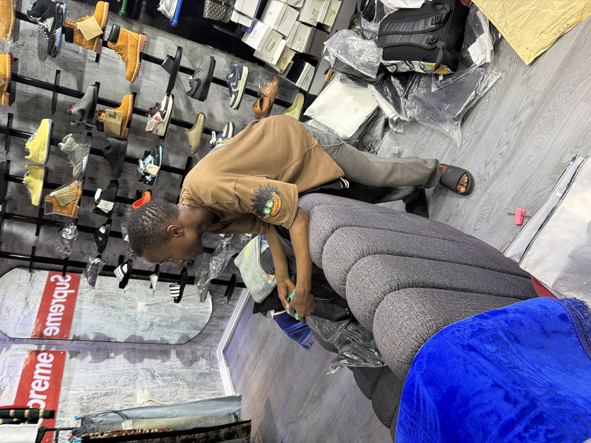 The image shows a young man playing a video game in a store located in Bukuru, Nigeria. The store is filled with shelves of shoes and clothing. The man is sitting on a plush grey sofa with a blue blanket draped over the armrest. He is wearing a brown t-shirt and grey sweatpants, with black slides on his feet. He is holding a green video game controller and appears to be focused on the game. In the background, shelves are stacked with various shoes, including Timberland boots, sneakers, and sandals. There are also boxes and bags of clothing and accessories. The store has a modern aesthetic with grey walls and wooden flooring. The overall atmosphere suggests a retail environment catering to young people.