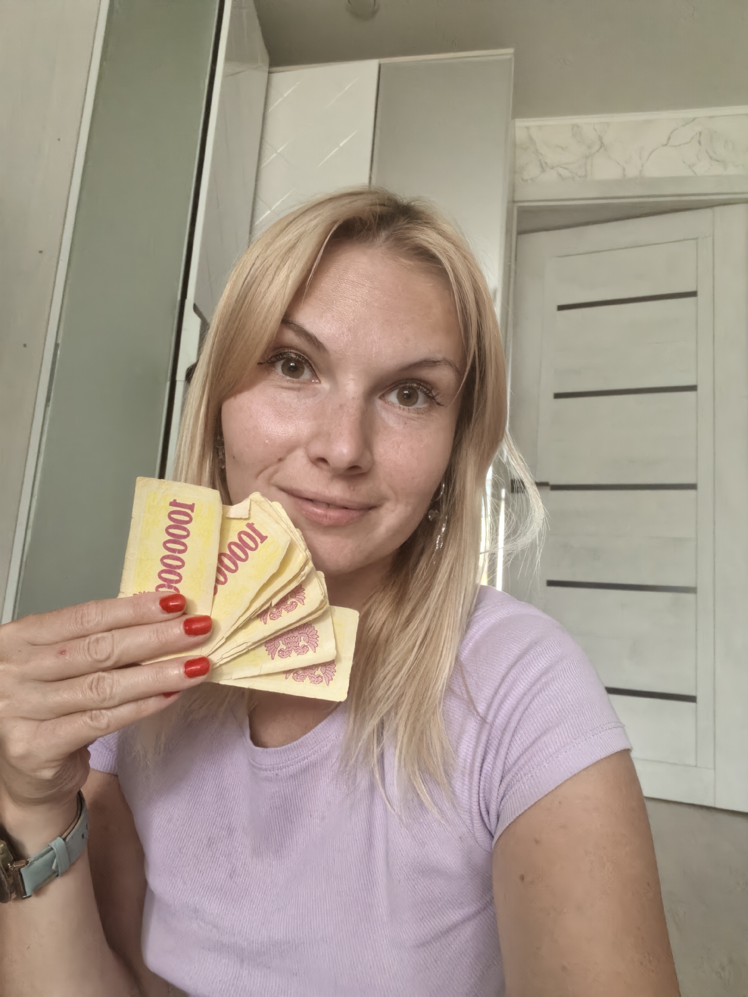 A woman with blonde hair and freckles is holding several yellow banknotes with red text that says "1000000" and a floral pattern. She is wearing a light purple t-shirt and a light blue watch. She is smiling and looking at the camera. The background appears to be an indoor setting, possibly a home, with white cabinets and a closed white door. The lighting suggests it is daytime. The provided context indicates the location as Sterlitamak, Russia.