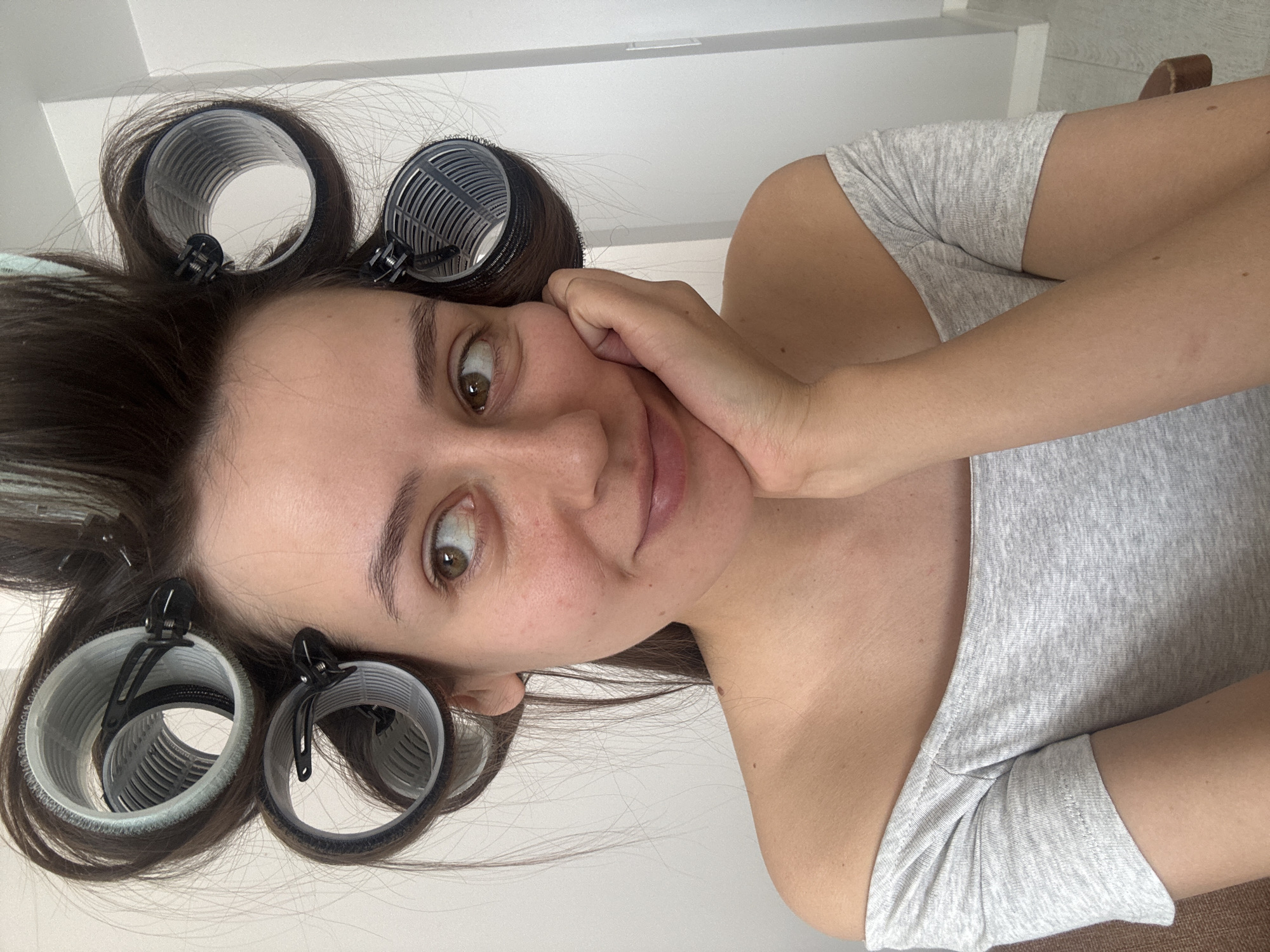 A young woman is lying on her side, facing the camera, with hair rollers in her hair. She is wearing a light grey off-the-shoulder top. Her left hand is near her face. The location appears to be indoors, potentially a bedroom or bathroom, with light-colored walls and furniture visible in the background. There are no indications that this image was taken in Krasnoyarsk, Russia.