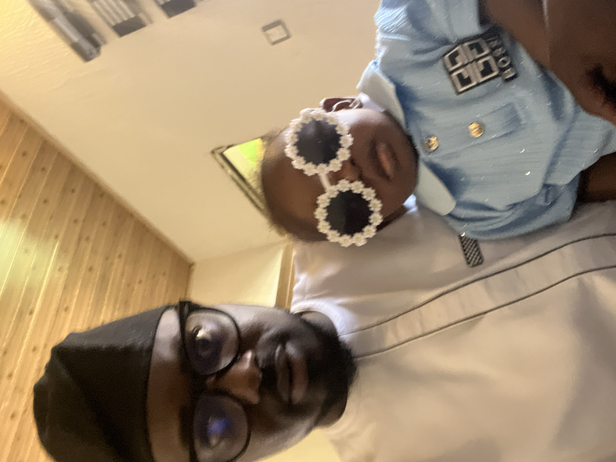 In the city of Barakin Akawo, Nigeria, a close-up, low-angle shot captures a man and a baby posing for a selfie. The man, wearing a black cap and glasses, is on the left, dressed in a white traditional outfit with dark trim. To his right, a baby is held, sporting novelty white floral sunglasses and a light blue collared shirt with gold buttons and a checkered "BOSS" logo. The background features a light-colored wall and a wooden ceiling.