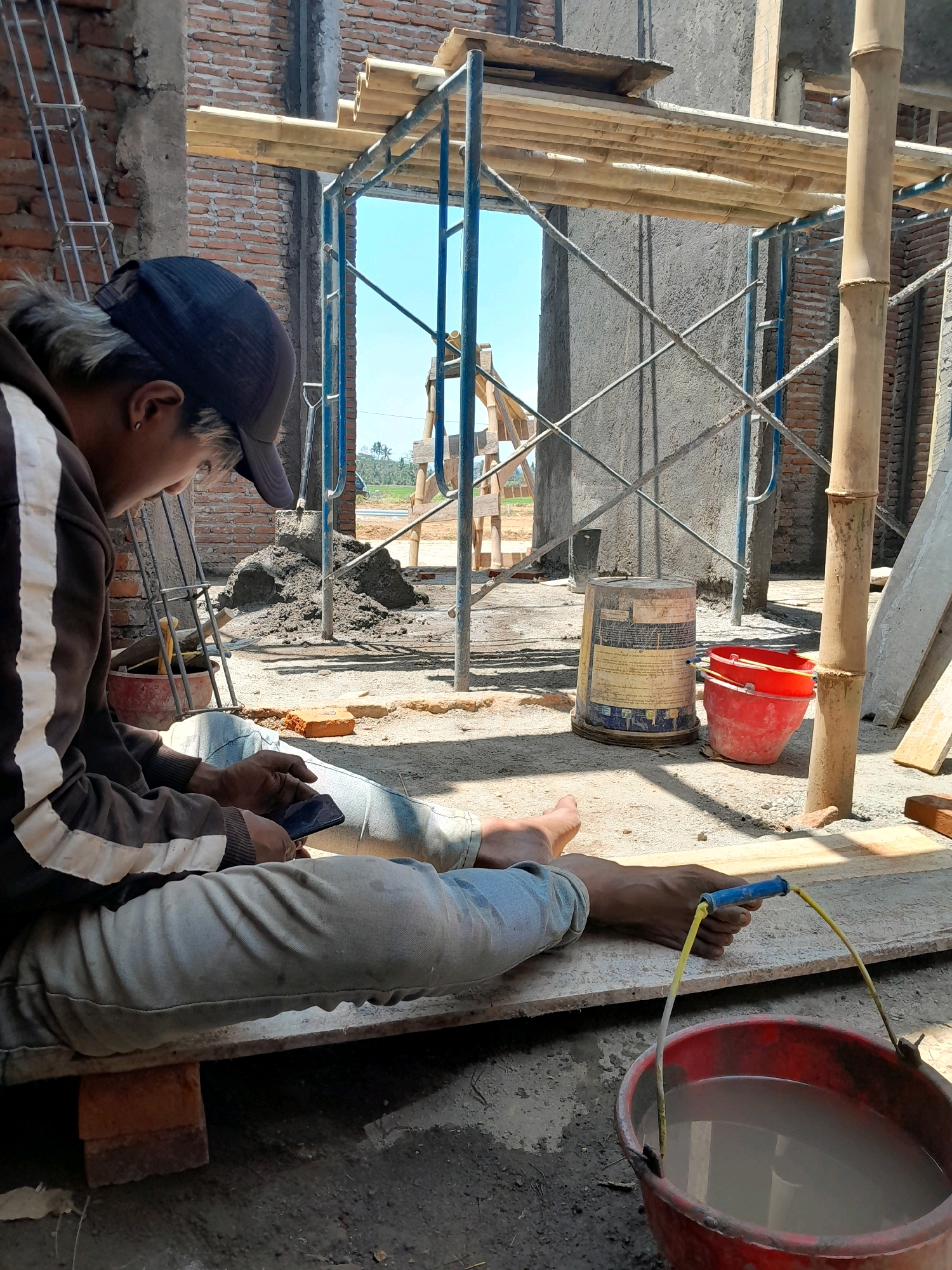 In Karangduren, Indonesia, a construction worker is sitting on a wooden plank, looking at his phone. He is wearing a dark blue baseball cap, a brown hooded jacket with white stripes, and light blue jeans. His bare feet are visible. In front of him, a red bucket with a yellow handle is filled with a light-colored liquid, possibly water or cement mix. Behind him, a pile of wet cement and construction materials, including scaffolding made of bamboo and metal poles, are visible. The background shows an unfinished brick building under construction. The scene is outdoors, with sunlight casting shadows on the ground.