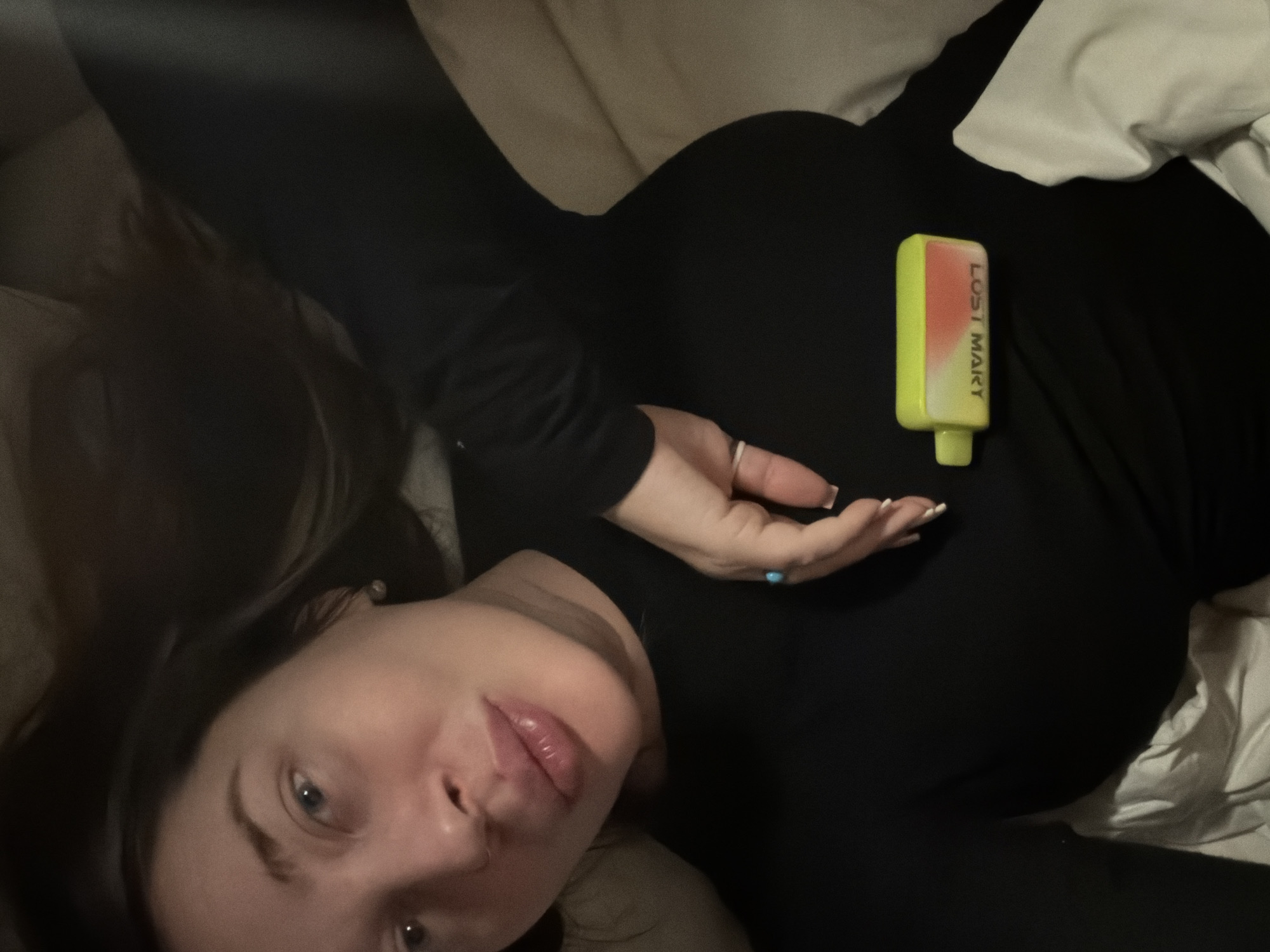 This indoor photograph, taken in Moscow, Russia, captures a female individual lying down, possibly on a bed or sofa, from a tilted, overhead perspective. The subject has long dark hair and blue eyes, and her face is positioned towards the camera with a neutral, relaxed expression. She is wearing a black long-sleeved top or dress.
Her left hand, adorned with a ring on the ring finger and a small blue accessory on another finger, is visible near her torso. Her nails appear manicured with white tips. Resting prominently on her chest is a two-toned vaping device, fluorescent yellow on one end and reddish-orange on the other. The brand name "LOST MARY" is clearly visible in white text on the device.
The background shows soft, light-colored bedding or pillows contrasting with darker areas, suggesting low ambient lighting, likely indicating evening or nighttime. The overall scene conveys a casual and intimate atmosphere.
