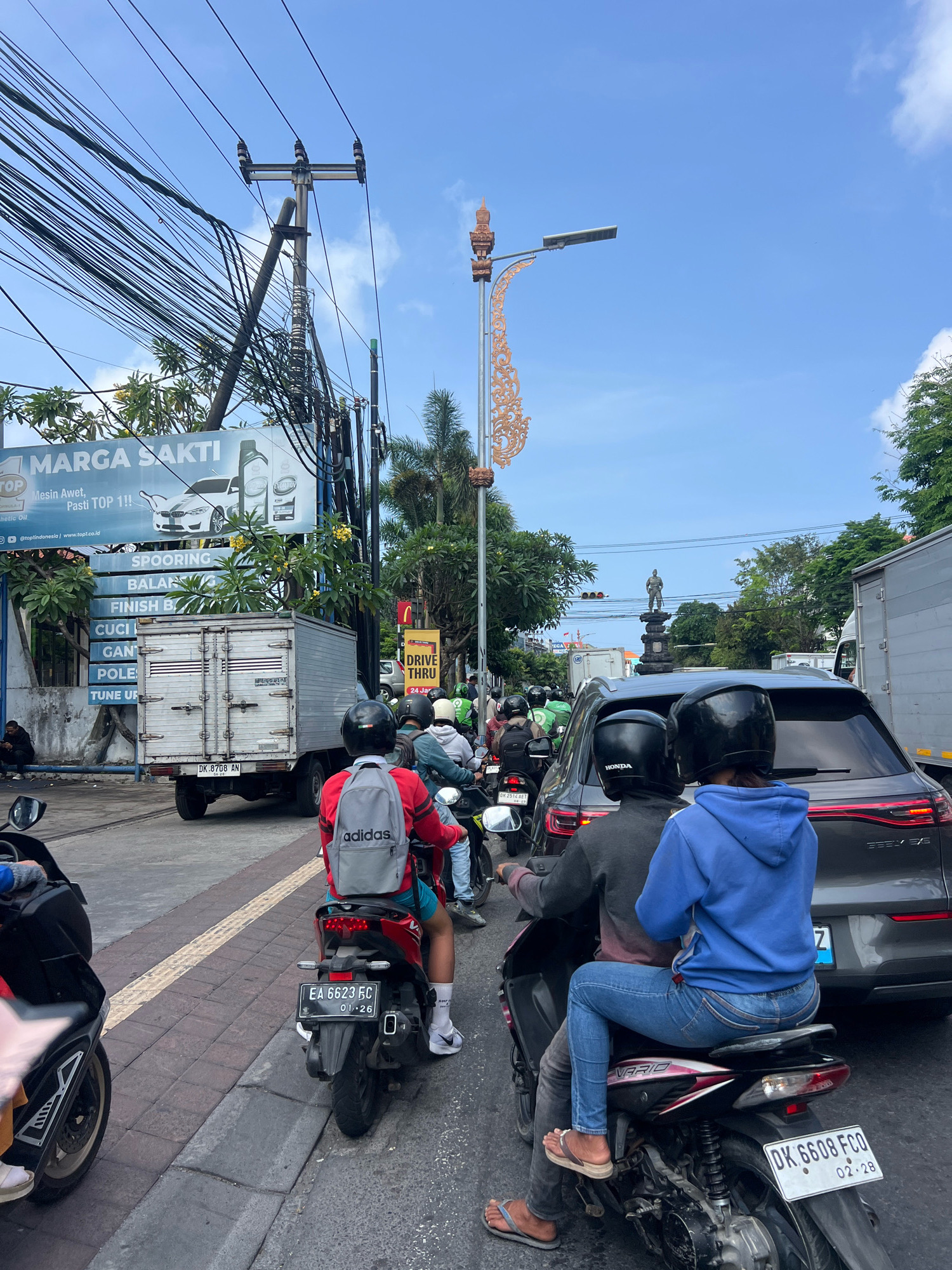 In Denpasar, Indonesia, a traffic jam is depicted on a city street under a bright blue sky with scattered clouds. Several motorbikes are stopped in a queue, with riders wearing helmets. One rider in the foreground wears a red jacket and a grey Adidas backpack, while the person behind them is wearing a blue hoodie and jeans. Behind the motorbikes, a grey SUV is visible, and further back, a large truck and other vehicles are present. A sign with the text "MARGA SAKTI" and "TOP" is displayed on a building to the left, along with a "DRIVE THRU" sign for McDonald's. Overhead, numerous electrical wires are strung between utility poles, and a decorative lamppost stands on the right side of the image. In the distance, a statue on a pedestal can be seen. The street is paved with bricks, and a yellow and white striped line marks the edge of the sidewalk.