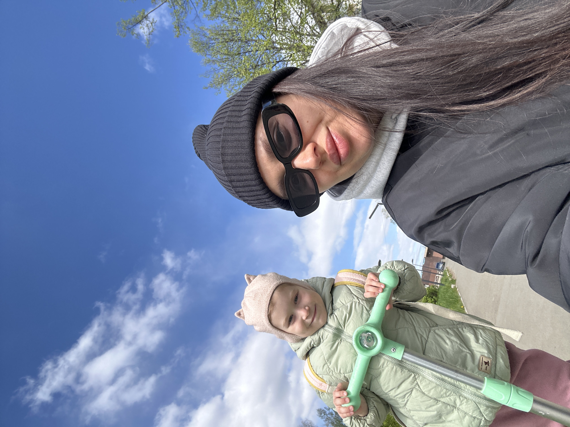 The image shows a selfie taken by a woman outdoors, with a child standing below her. The woman is wearing a black beanie, sunglasses, a grey hooded sweatshirt, and a black jacket. She has long dark hair. The child is wearing a light pink hat with ears, a light green quilted jacket with yellow stripes, and pink pants. The child is holding onto the green handlebars of a scooter. The background includes a blue sky with clouds, trees with green leaves, and a sidewalk or road. The location is in the city of Chaikovsky, Russia.