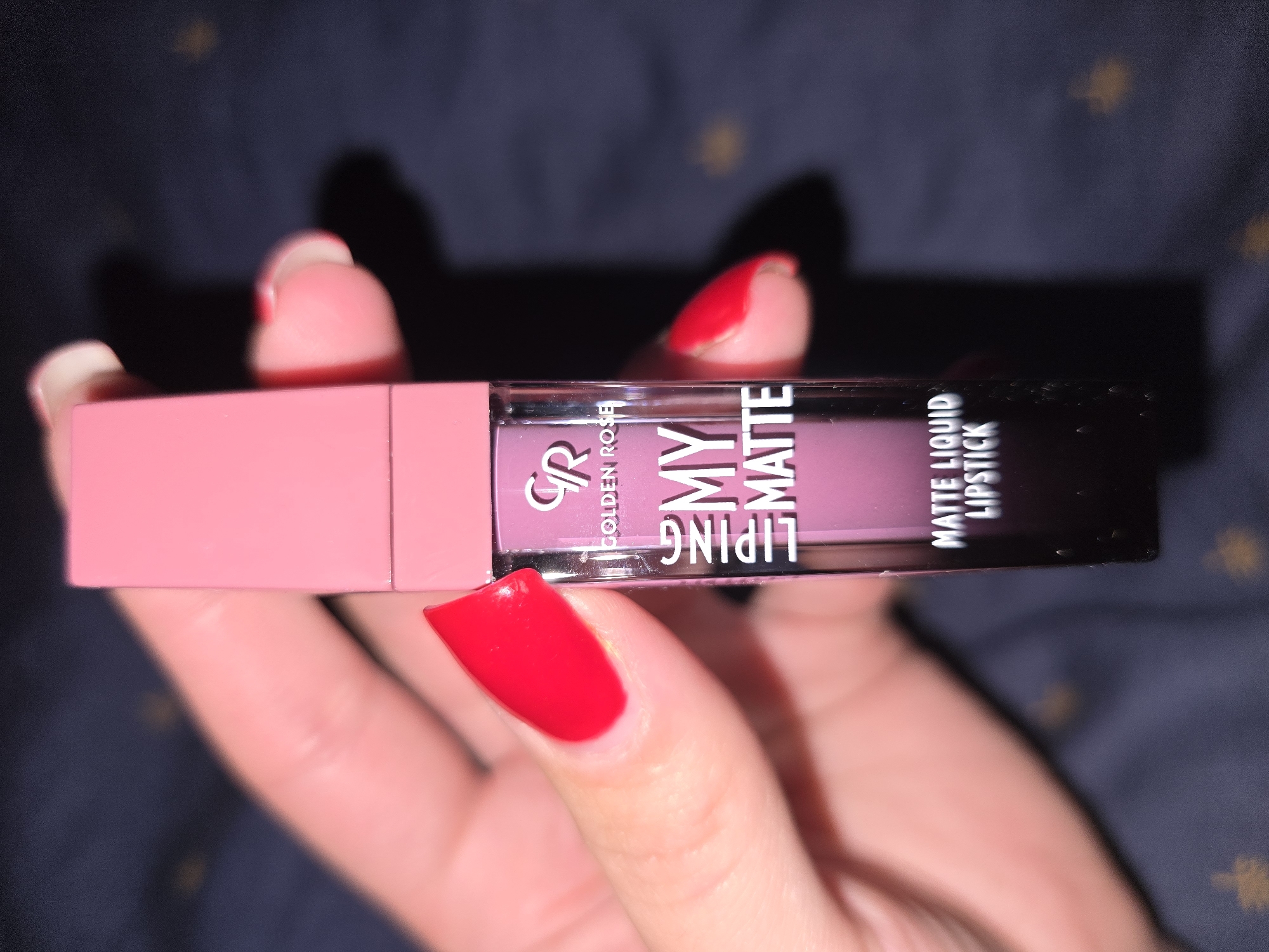 A person's hand, with bright red nail polish, is holding a pink and clear tube of Golden Rose "My Matte" lip liquid lipstick. The product is angled towards the viewer, with the clear tube showing the purple lipstick inside. The background is a dark, textured surface with some blurry yellowish circular shapes, possibly lights or decorations. The lighting suggests it might be indoors, potentially at night. The hand's fingers are visible, adding a human element to the product shot. The text "GOLDEN ROSE", "MY", "MATTE", and "LIPSTICK" are clearly legible on the packaging. The setting appears to be a close-up, personal shot, focusing on the makeup product.