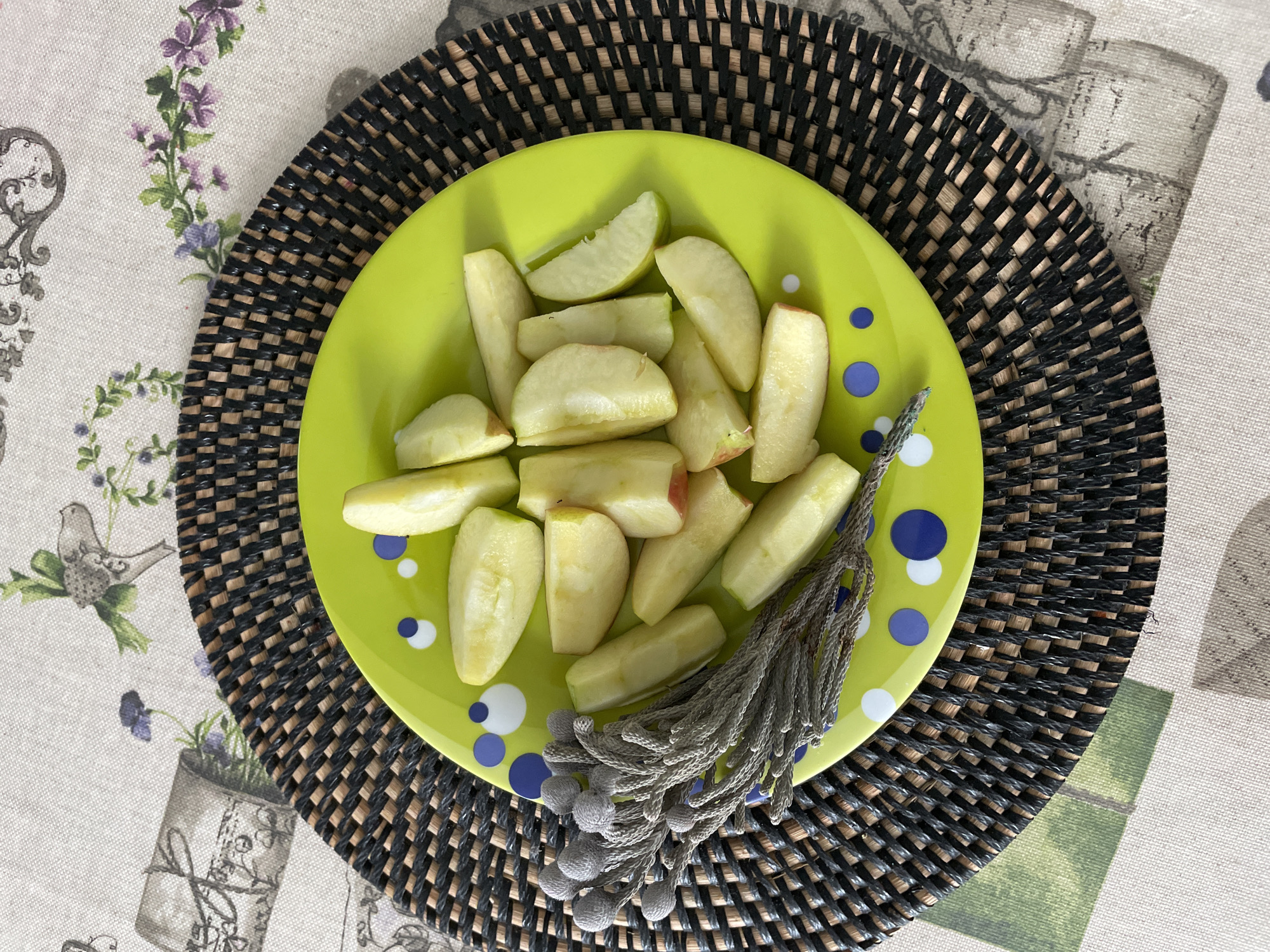 A top-down shot reveals a plate of sliced apples on a black woven placemat. The light green plate, speckled with blue and white circles, holds approximately fifteen apple slices, their pale flesh tinged with hints of green and red peel. A decorative gray tassel lies adjacent to the apples. The placemat sits atop a patterned tablecloth featuring illustrations of flowers, birds, and vases. The scene is likely located in the Лесной Пейзаж Кп (шарапово) area of Russia, based on the photo's origin.