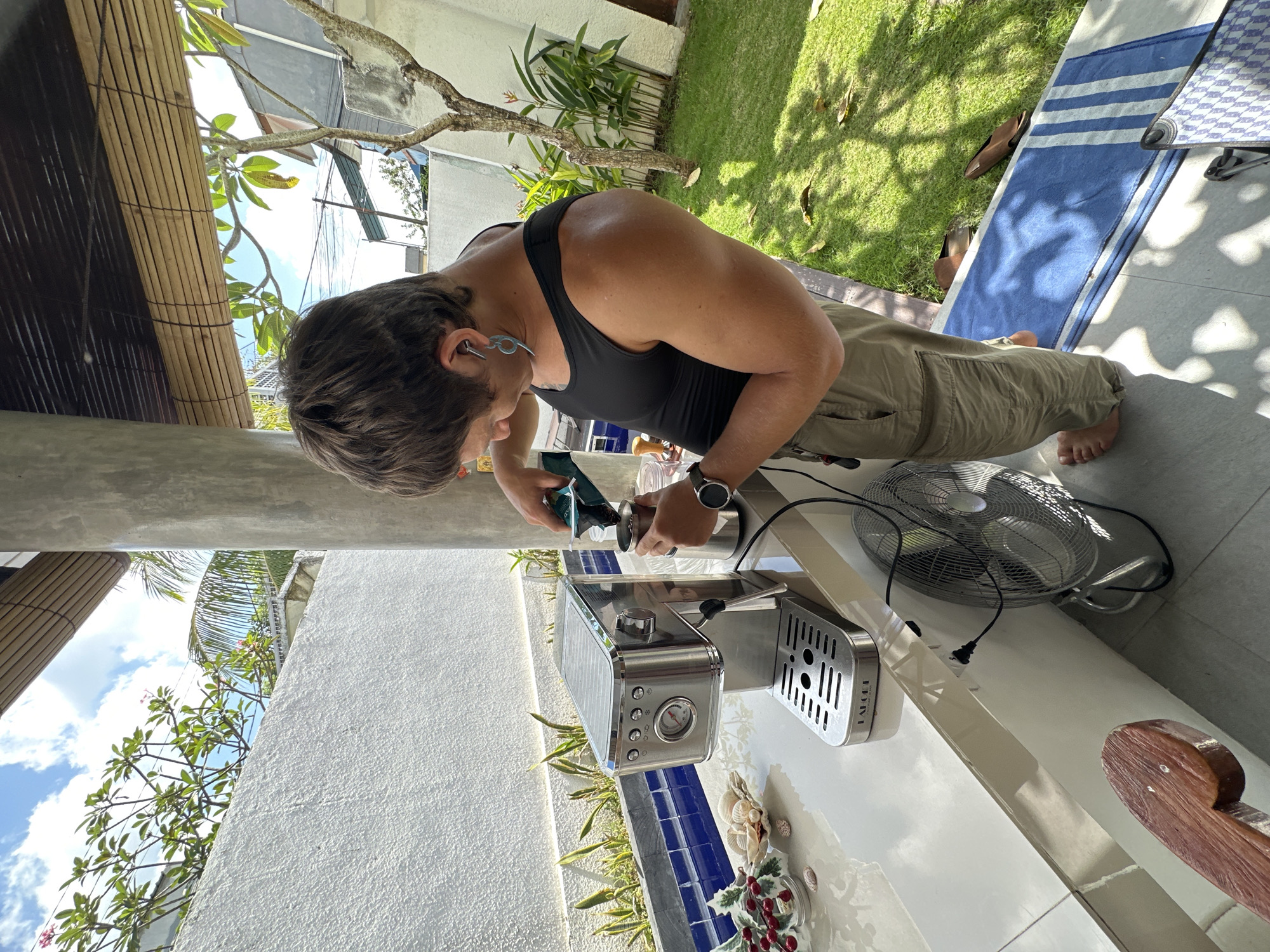 A person stands outdoors near a white wall and lush green grass, preparing coffee. They are wearing a black tank top and khaki cargo pants, with one foot bare and the other partially obscured by a blue and white striped mat on the ground. The person is actively engaged in making coffee, pouring grounds from a bag into a metal container held by their left hand, while their right hand operates a silver espresso machine. The machine sits on a white countertop, adorned with a shell-filled dish and a small decorative arrangement of faux berries and leaves. A black electric fan is positioned on the floor beneath the countertop, plugged into an outlet. The background reveals tropical vegetation, a concrete pillar, and a bamboo-slatted covering overhead, suggesting a patio or outdoor living space in North Kuta, Indonesia. The sky above is blue with scattered white clouds.
