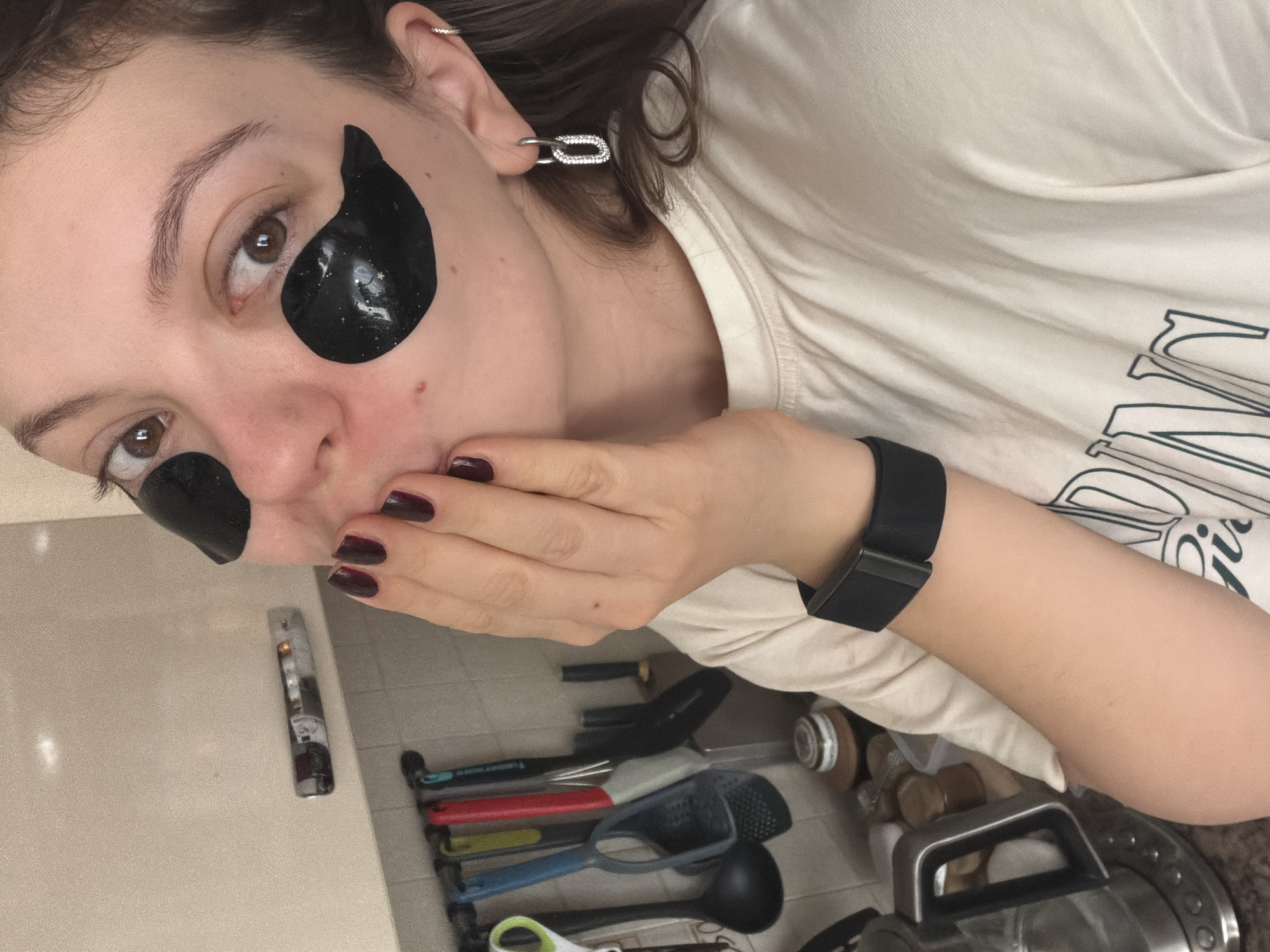 A close-up, tilted portrait captures a young woman engaged in a self-care moment within what appears to be a kitchen or utility area in Khimki, Russia. She looks directly towards the camera with a neutral expression, her dark brown eyes prominent.
The woman has fair skin and dark brown hair. She is wearing black, shimmering hydrogel under-eye patches. Her left hand is raised to her mouth, with dark red nail polish visible on her short nails, covering part of her chin and lower lip. On her left wrist, she wears a black smartwatch or fitness tracker. Her ear is adorned with two silver-toned earrings: a small hoop or cuff near the helix and a more prominent link-shaped earring, embellished with small gems, dangling from the lobe.
She is dressed in a light cream or off-white t-shirt, on which dark, stylized text is partially visible. The letters "N" and "O" can be discerned as part of an unknown word or logo.
The background suggests an indoor setting. A light-colored countertop is visible on the left. On the right, a collection of kitchen utensils with colorful handles (red, grey, blue) – including spatulas, a whisk, and a black ladle – are arranged in what appears to be a drawer or container. A small, clear spray bottle rests on the countertop near her hand. A large, metallic kitchen appliance, possibly an air fryer or similar, is partially visible in the lower right corner. The lighting is soft and ambient, suggesting an indoor setting without strong natural light. The overall mood is calm and personal.