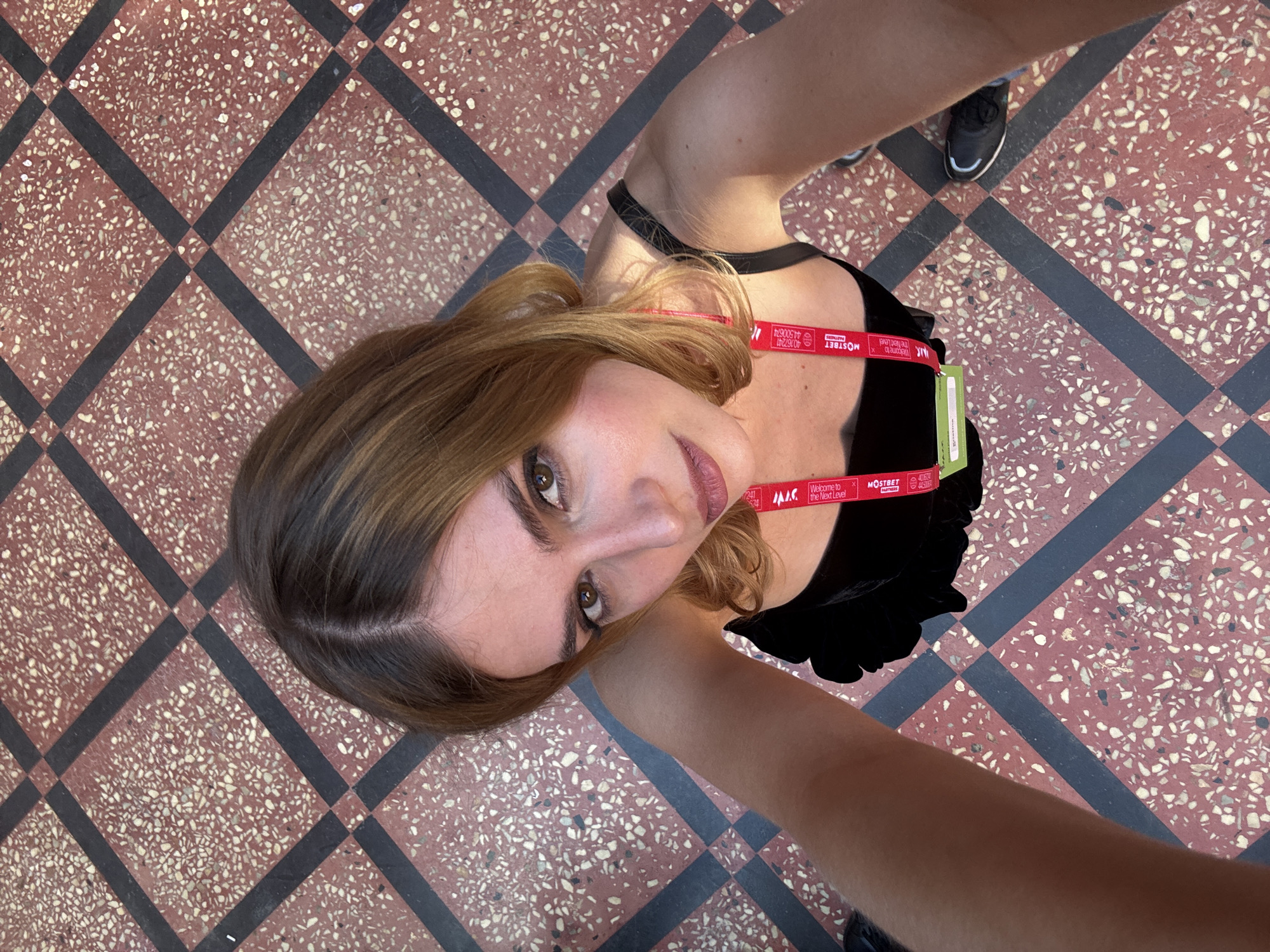 A person is photographed from a low-angle perspective against a background of red and white terrazzo flooring with black gridlines, suggesting a public interior space. The individual is wearing a black top and a lanyard with a red strap and a green tag around their neck. The lanyard has the text "Welcome to the Next Level" and "MOSTBET" printed on it. The photograph captures part of the person's legs, clad in dark-colored shoes, visible towards the periphery. The surroundings are well-lit. The image was taken at the Moscow Cinema in Yerevan, Armenia.