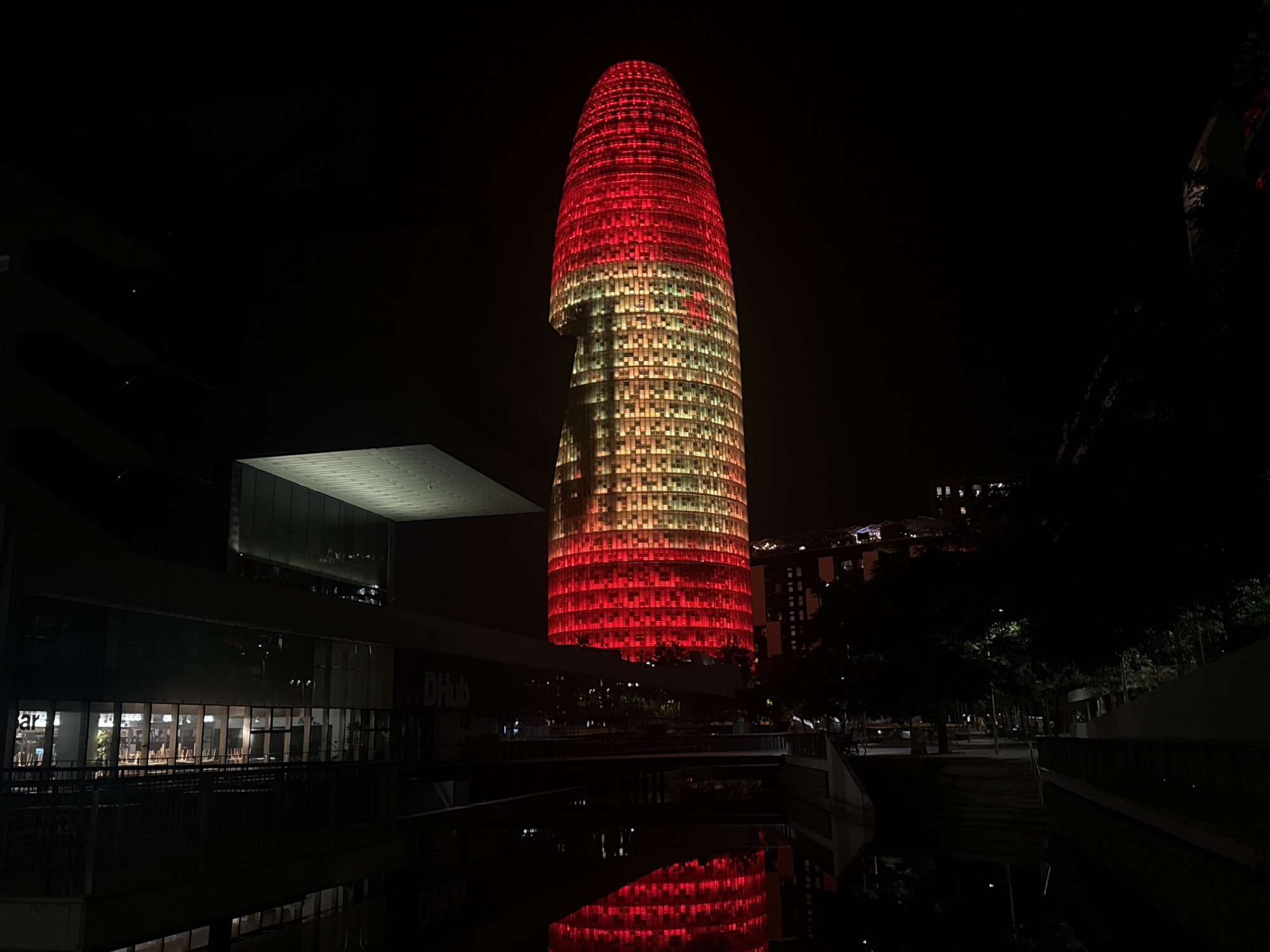 The image shows the Torre Glòries, a skyscraper located in Barcelona, Spain, at night. The building is illuminated with red and yellow lights, creating a striking visual effect. In the foreground, a body of water reflects the illuminated skyscraper and the surrounding dark environment. To the left of the skyscraper, a modern building with a flat roof and glass facade is visible. The surrounding area is dark, with some trees and other buildings faintly visible in the background. There are no people or animals clearly visible in the image. The scene appears to be an urban landscape at night, with the illuminated skyscraper being the central focus.