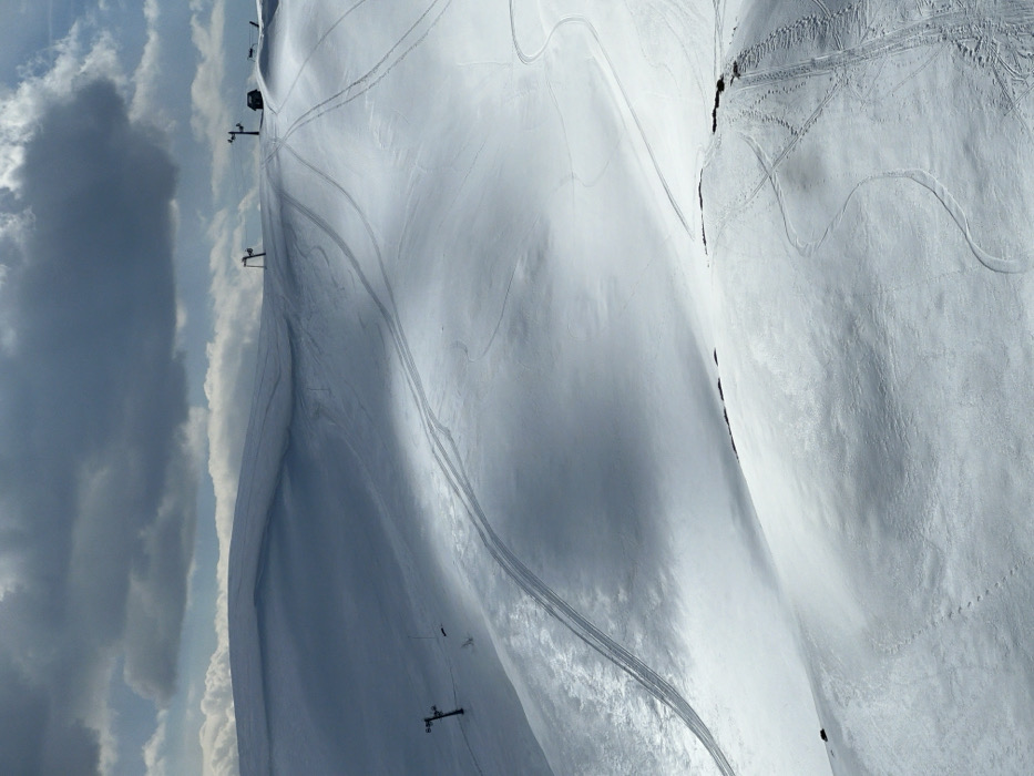 This image depicts a snow-covered mountainous landscape in Katun Bačko brdo, Montenegro, under a partially overcast sky. The scene is dominated by undulating terrain covered in white snow, marked with numerous tracks. Several ski lift poles are visible, primarily along the upper ridge of the mountains, indicating an operational or recently used ski area. Two distinct types of tracks are observable: narrower, winding tracks consistent with skis or snowboards, and wider, straighter tracks, possibly from snow groomers or snowmobiles, traversing the slopes. A small, dark structure, potentially a service building or shelter, is situated near one of the uppermost ski lift poles on the right side of the image. Sunlight intermittently illuminates sections of the snow, creating contrasting areas of bright white and cooler shadows. No individuals are visible. The sky features dark grey clouds interspersed with brighter patches.