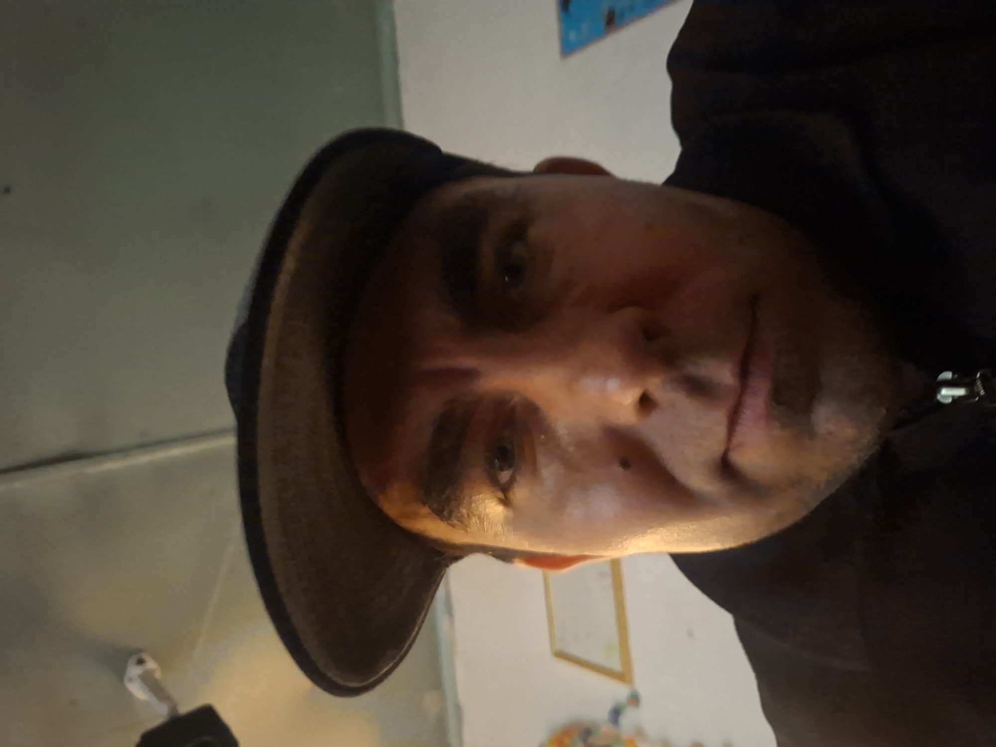 A first-person perspective selfie captures a man wearing a black jacket and a dark baseball cap. His face, partially illuminated by warm lighting, is the central focus. The background is indistinct, with a wall that appears to be painted a light color. A framed picture with a golden border is visible on the wall to the left of the man. A faint white string hangs from the ceiling in the upper left corner of the image, attached to an object that is out of frame.