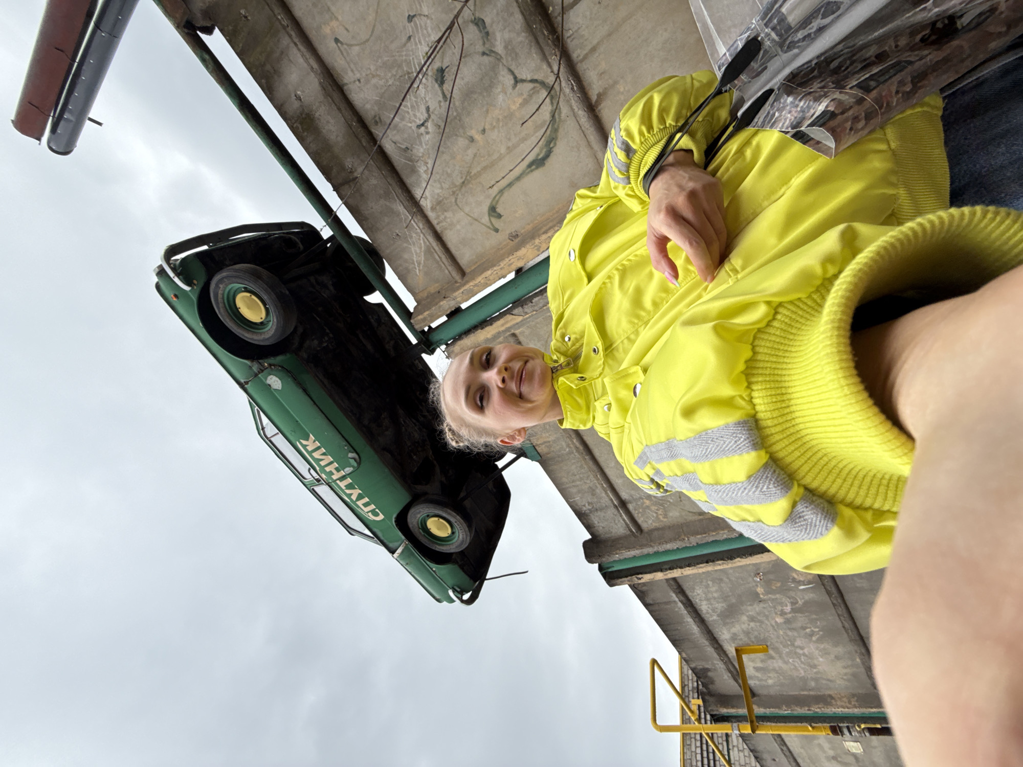 The image captures a unique outdoor scene in Perm, Russia, featuring a woman taking a selfie with a vintage green car art installation. The woman, with blonde hair pulled back, smiles pleasantly at the camera. She is wearing a bright, high-visibility yellow jacket with reflective gray stripes on the sleeves, the ribbed cuff of which is prominent in the foreground.
Above and behind her, a dark green, older model car is mounted in an unusual inverted position against the underside of a weathered concrete overhead structure, possibly an overpass or part of a building. The car's wheels feature distinctive yellow hubs. The sky visible in the background is overcast, indicating a cloudy daytime.
A significant detail is the Cyrillic text clearly visible on the side of the car, which spells out "СПУТНИК" (Sputnik). This suggests the car is either a model named Sputnik, or part of an art installation referencing the Soviet space program or a similar theme. The concrete structure shows signs of age, with some visible staining or growth. Green metallic beams are also integrated into the structure. The scene presents an intriguing blend of urban environment and public art.
