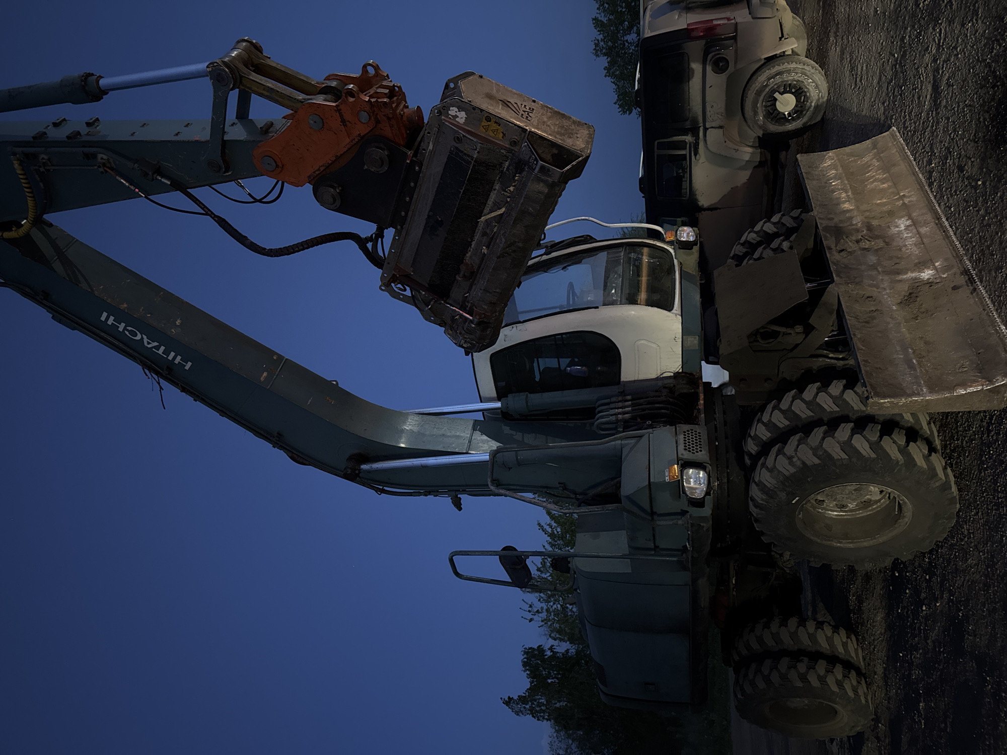 The image shows a Hitachi excavator parked at night in an unknown location. The excavator has a grapple attachment on its arm. Beside the excavator is a truck with a partially visible plow. The surrounding environment appears to be a parking lot or storage area.