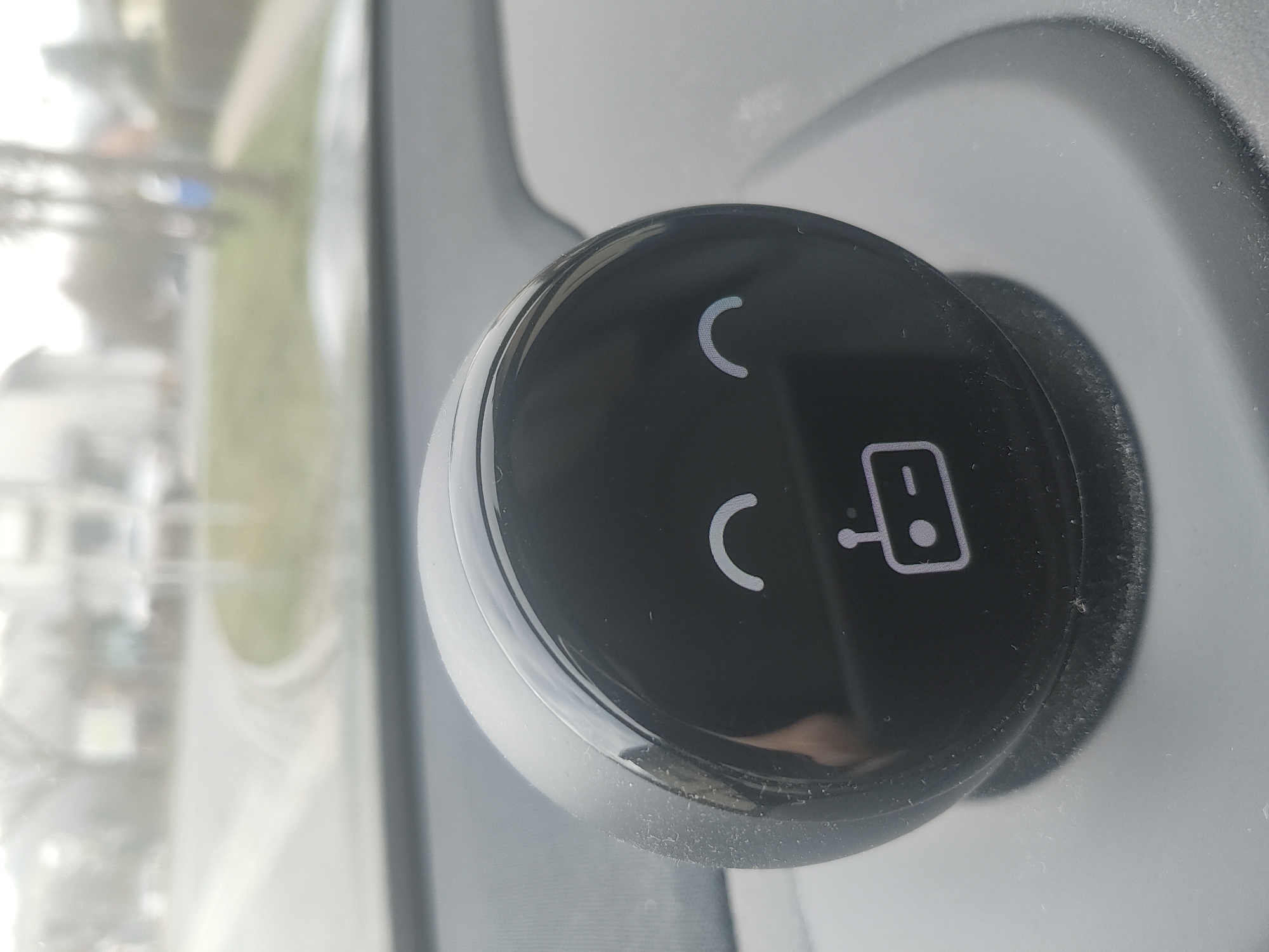 The image displays a close-up of a black, circular, glossy control knob or button, likely part of an electronic device or vehicle interior. The knob is positioned against a light gray surface, which forms the immediate background.
On the face of the black knob, three white symbols are visible. On the left side, two identical crescent-shaped symbols are stacked vertically. These resemble either open parentheses or the letter 'C'. On the right side, there is a symbol depicting a rectangular card with a circular element (suggesting a chip) being inserted into a narrow slot. The card is oriented horizontally, and the slot extends to its right. These symbols appear illuminated or are made from a reflective white material.
The black surface of the knob is highly reflective, showing faint, blurry reflections of the surroundings, including a partial reflection of what might be the person taking the photo at the bottom center. Some dust particles are visible on the gray surface surrounding the knob and along its edges.
Beyond the immediate interior, a blurred outdoor scene is visible through what appears to be a window. This background shows indistinct shapes of green (possibly grass or trees) and lighter, hazy areas, suggesting a bright, possibly daytime setting with diffuse light. The overall impression is that of an interior setting, likely a vehicle, with a view to the outside.