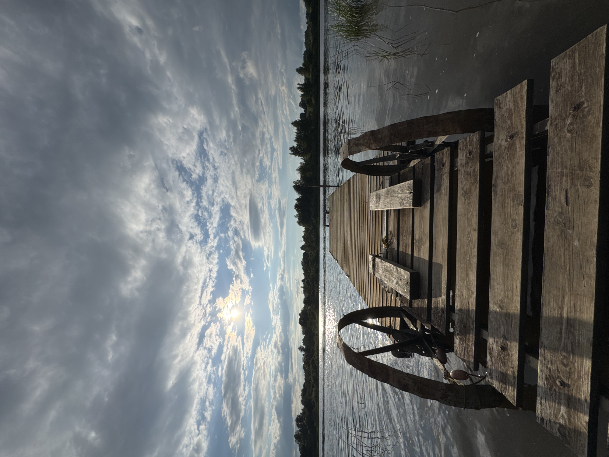 The image depicts a wooden pier extending into a calm body of water, likely a lake or river, in Sitno, Russia. The sky above is a mix of dramatic grey clouds with patches of bright sunlight breaking through, creating reflections on the water's surface. The pier, made of weathered wooden planks, has two rustic metal wheels attached to its sides near the entrance. These wheels appear to be part of a mechanism, possibly for launching or retrieving a boat. The far bank of the water is lined with a dense forest. The scene is serene and natural, with no people visible. The overall impression is one of a quiet, somewhat remote waterfront location.