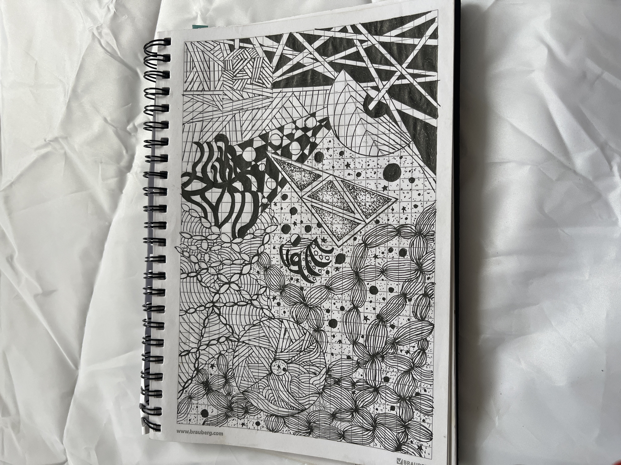 A black and white sketch fills a page in a spiral-bound notebook. The drawing is abstract, composed of various geometric patterns, lines, and shaded areas. The notebook is placed on a white, wrinkled surface, possibly fabric. The image does not depict any location in Krasnyye Barrikady, Russia, or any identifiable city elements.
