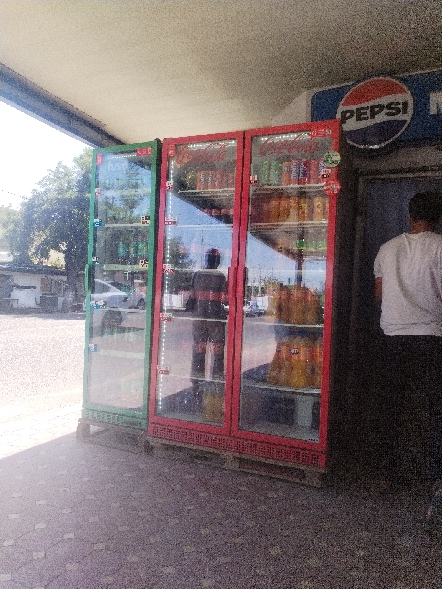Two commercial refrigerators are visible, one green and one red, filled with bottled beverages. The green refrigerator displays a "fuse" logo at the top and pricing labels on its glass doors. The red refrigerator, positioned to the right of the green one, is filled with various sodas, including Coca-Cola and Fanta, also with visible pricing labels. Behind the refrigerators, a Pepsi sign is mounted on a blue background. A person in a white t-shirt and dark pants stands to the right of the red refrigerator, facing away from the camera. The scene appears to be an outdoor market or roadside stall. The ground is paved with interlocking grey tiles. In the background, there are trees, a building with a white facade, and parked vehicles, including a white car and a white truck. The environment suggests a warm, sunny day. The location is identified as Tashkent, Uzbekistan.