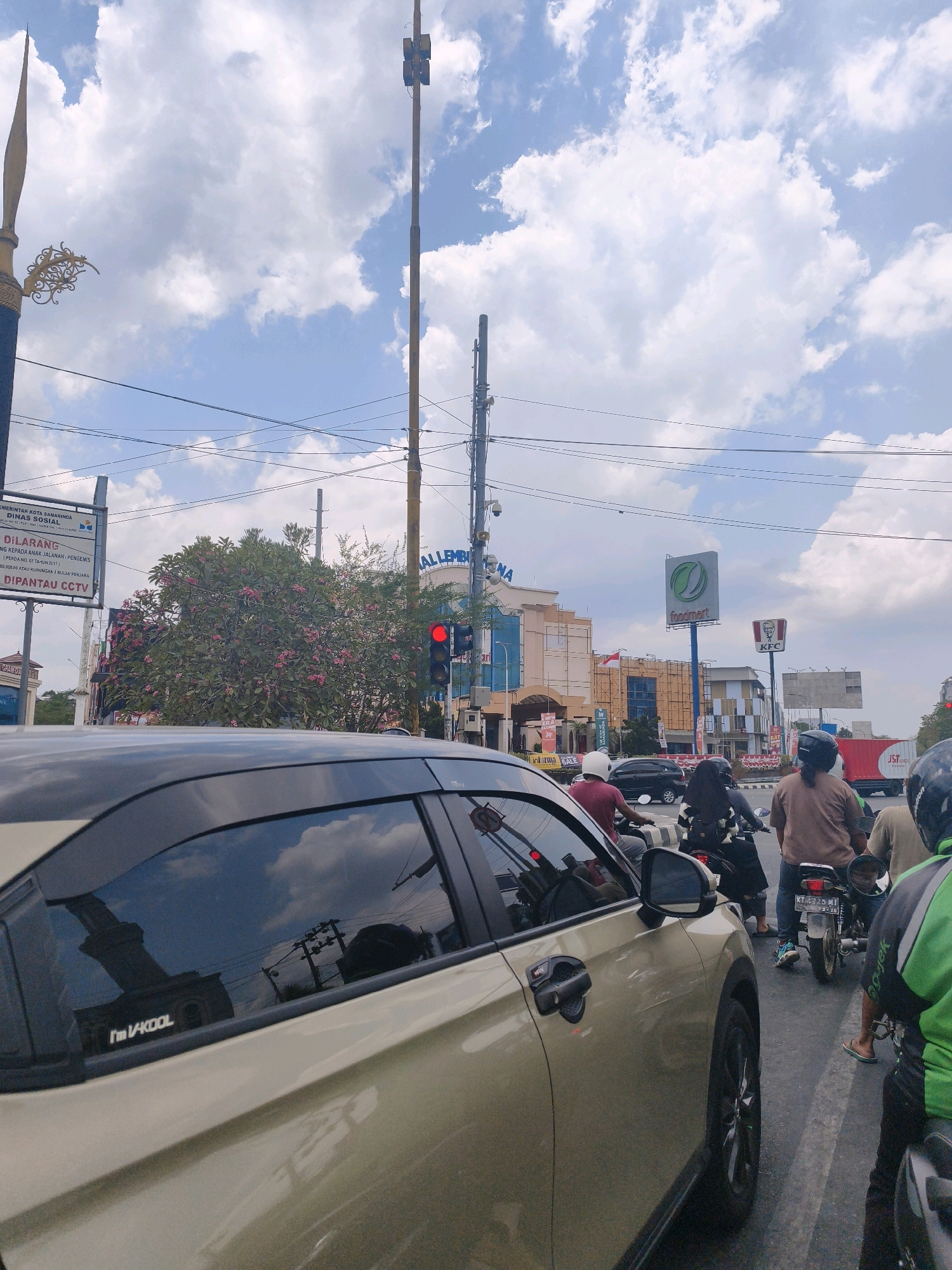 The image depicts a street scene in Samarinda, Indonesia, likely during the daytime under a partly cloudy sky. A tan-colored car is prominently featured in the foreground, with its side and rear window visible. Reflected in the car's window is a building and overhead wires. Several motorcycles and their riders are also visible in traffic, moving along the asphalt road.
In the background, there are various buildings, including a KFC restaurant and a building with a "foodmart" sign. A traffic light is illuminated red, indicating a stop. There are also several utility poles with numerous wires crisscrossing the sky. A street sign displaying information from the "PEMERINTAH KOTA SAMARINDA DINAS SOSIAL" (Government of Samarinda City, Social Services) is visible on the left, along with a warning about surveillance cameras. A tree with pink flowers is also present. The overall impression is of a busy urban intersection.