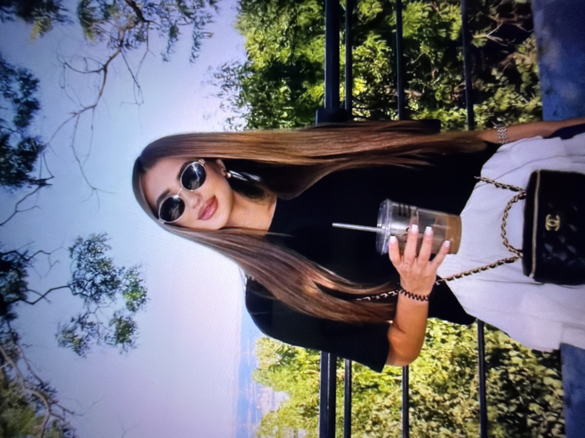 In Beșalma, Moldova, a young woman stands on a balcony, holding a beverage and a handbag. She is dressed in a black top and a white ruffled skirt, accessorized with sunglasses, earrings, and a bracelet. Her long, brown hair cascades down her shoulders. The background features lush greenery and a distant cityscape, suggesting a pleasant day with clear skies.