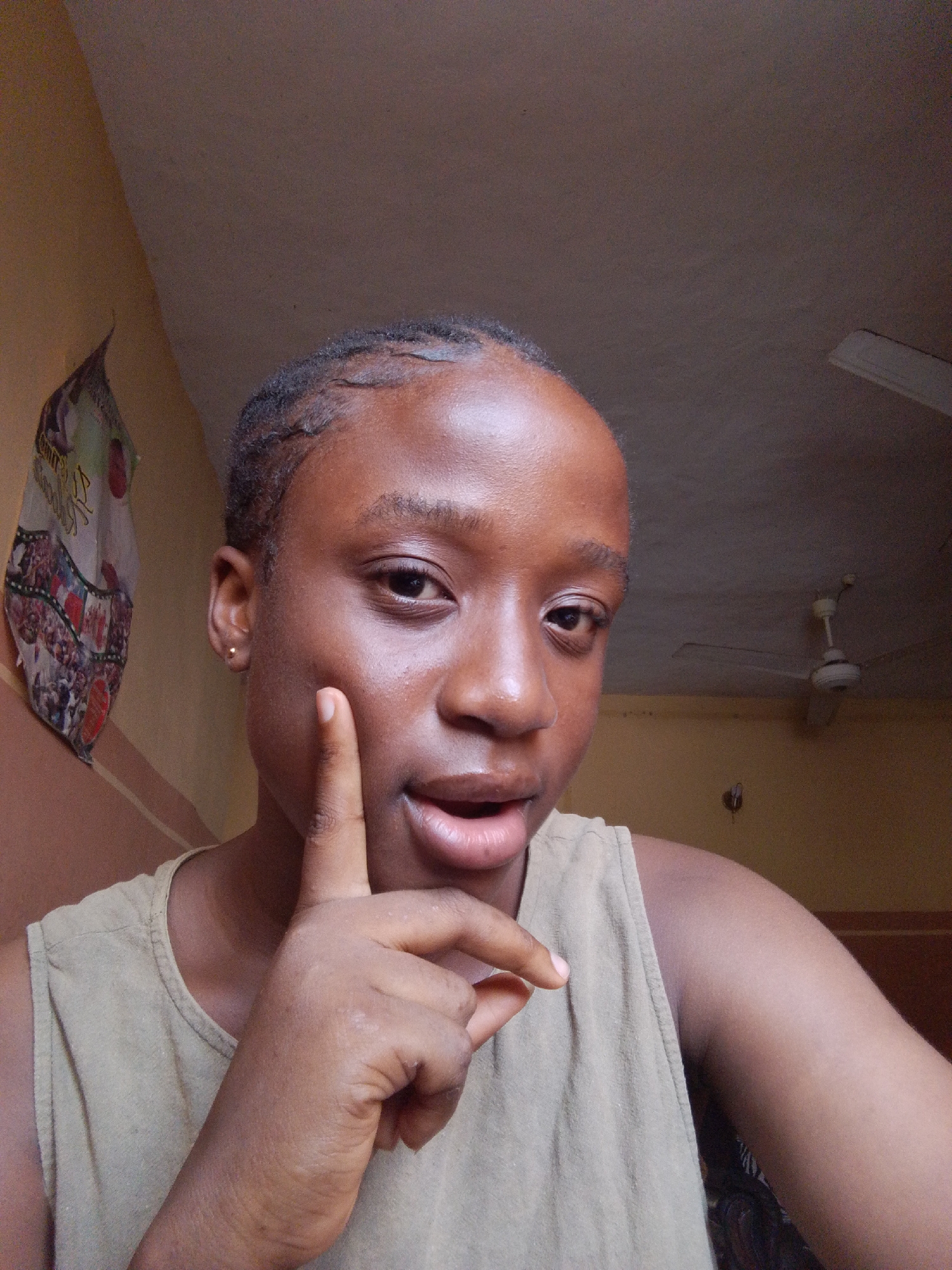 A person is shown in a close-up, indoor shot. The person has brown skin, short, dark hair styled close to the head, and is wearing a tan sleeveless top. The person's right hand is near their face, with the index finger extended and touching the cheek. The person's mouth is slightly open. In the background, the walls are painted in various shades of yellow and beige. A decorative item hangs on the wall to the left. A ceiling fan is visible in the upper right corner of the frame. The location is Unknown, Unknown.