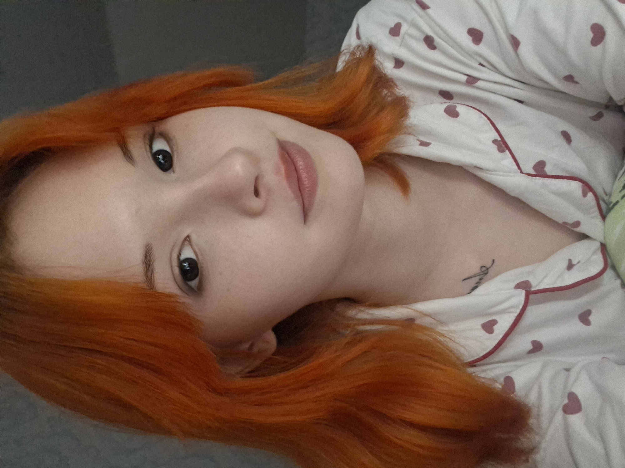 The image features a young woman with striking, coppery-orange hair, lying down and looking directly into the camera. Her head is tilted, giving a relaxed and engaging pose. She has fair skin, dark eyes with visible reflections, light eyebrows, and soft pink lips that curve into a subtle smile.
She is wearing white pajamas patterned with numerous small, light pink hearts and trimmed with thin red piping along the edges of the garment, particularly visible on the collar and placket. Her vibrant hair spreads around her head, contrasting with a dark, muted background, possibly a wall or headboard.
A small, black script tattoo is visible on her upper chest, near her collarbone, partially obscured by the neckline of her pajama top. The exact text of the tattoo is not clearly discernible from the image. The setting appears to be indoors, suggesting a comfortable environment like a bedroom, emphasized by her casual attire and reclining posture. A hint of another fabric, possibly a green and white stripe, is visible on the far right edge of the frame. The overall atmosphere is intimate and calm.