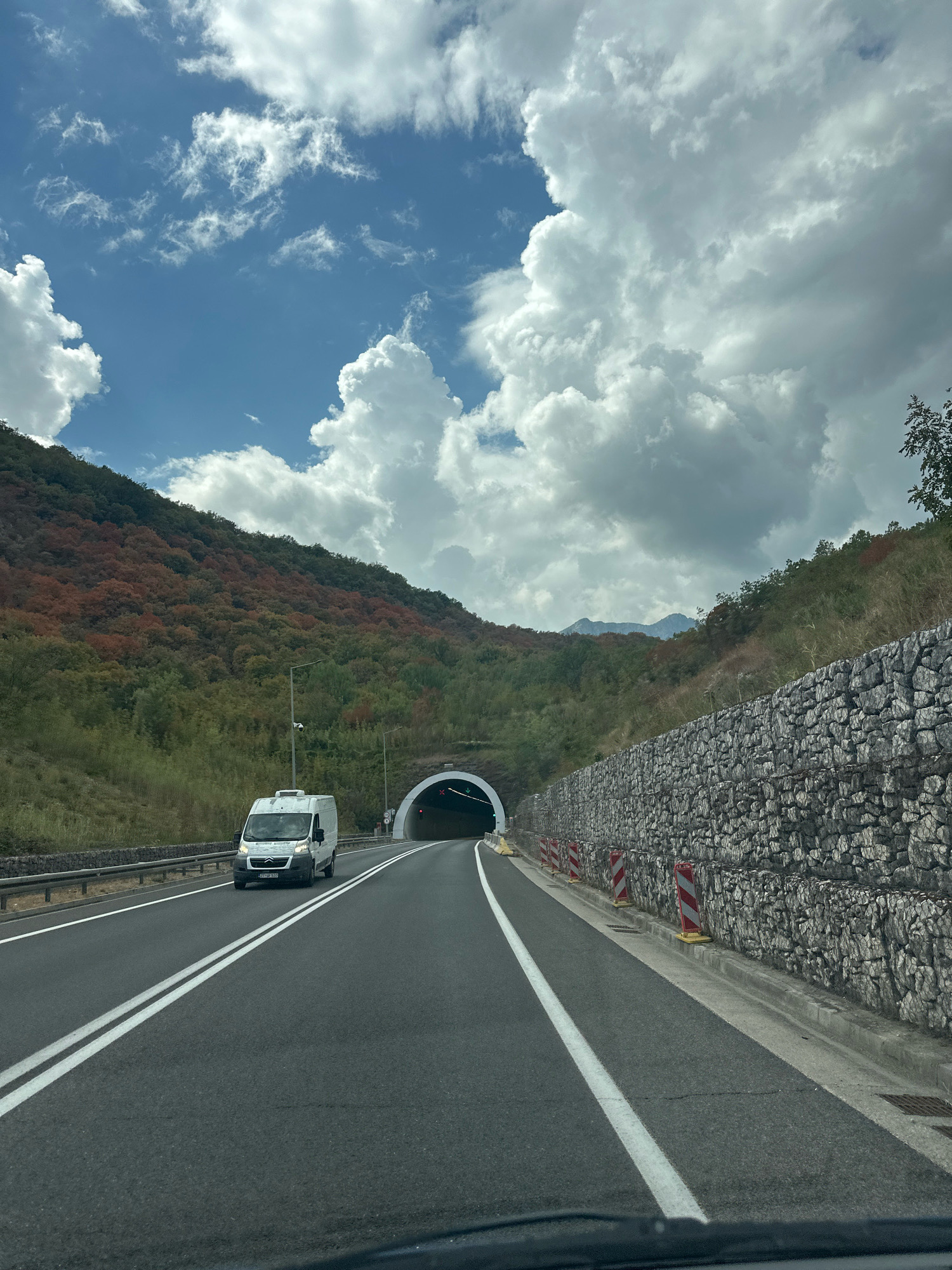 A white van travels on a two-lane asphalt road towards a tunnel entrance. The tunnel is illuminated and has a red traffic light displayed, indicating it is closed. The road is bordered by a concrete barrier on the right, which is reinforced with a gabion wall constructed of stacked stones. On the left, the road is also bordered by a metal guardrail, behind which are green trees and a hillside covered with vegetation. The hillside displays patches of autumnal colors, with trees showing shades of orange and red among the green. The sky above is a mix of blue and white, with large, fluffy cumulus clouds scattered across it. The scene appears to be set in a mountainous or hilly region, likely a highway or main road leading to or from a tunnel. The location is identified as Sotonići, Montenegro.