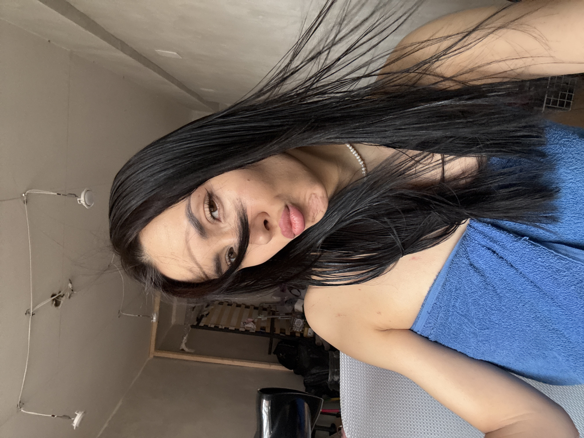 A woman with long, dark hair is seen from a close-up, canted angle. She is wearing a blue towel and a pearl necklace. The background shows a room with exposed electrical wiring in the ceiling, a framed structure (possibly a bed frame), and miscellaneous objects. The location in the city of Votkinsk, Russia cannot be determined from the image.