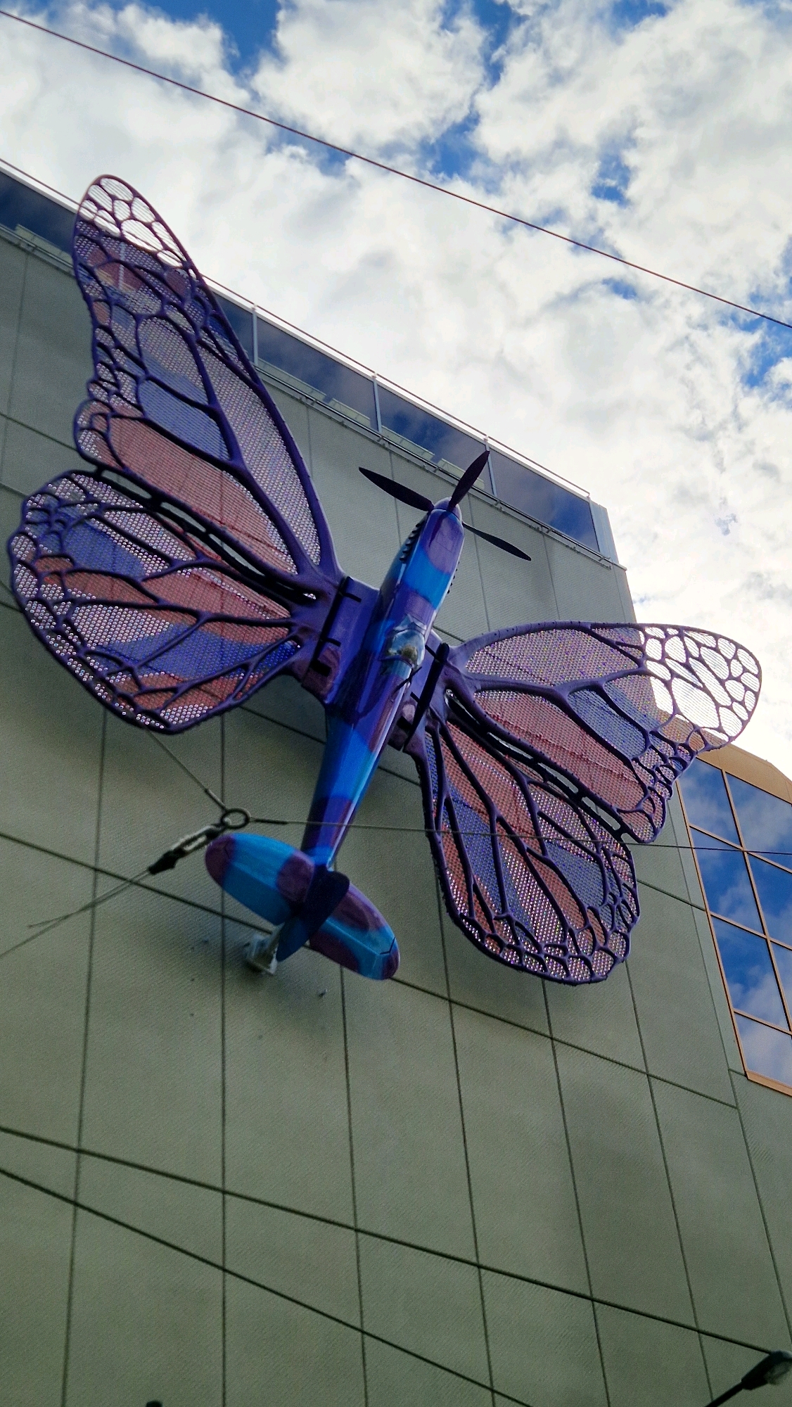 An art installation depicting an airplane with butterfly wings is mounted on the exterior wall of a modern building. The airplane is painted in shades of blue and purple with pink accents on the wings, which are designed with a mesh-like pattern resembling butterfly wings. The building has a neutral-colored facade and large, reflective windows. The sky above is partly cloudy, with white clouds against a blue background. The installation is suspended by wires, one of which is visible attaching to a metal bracket on the wall. This artwork is located in Prague, Czechia, specifically at the Dancing House, also known as Fred and Ginger, on Rašínovo nábřeží.