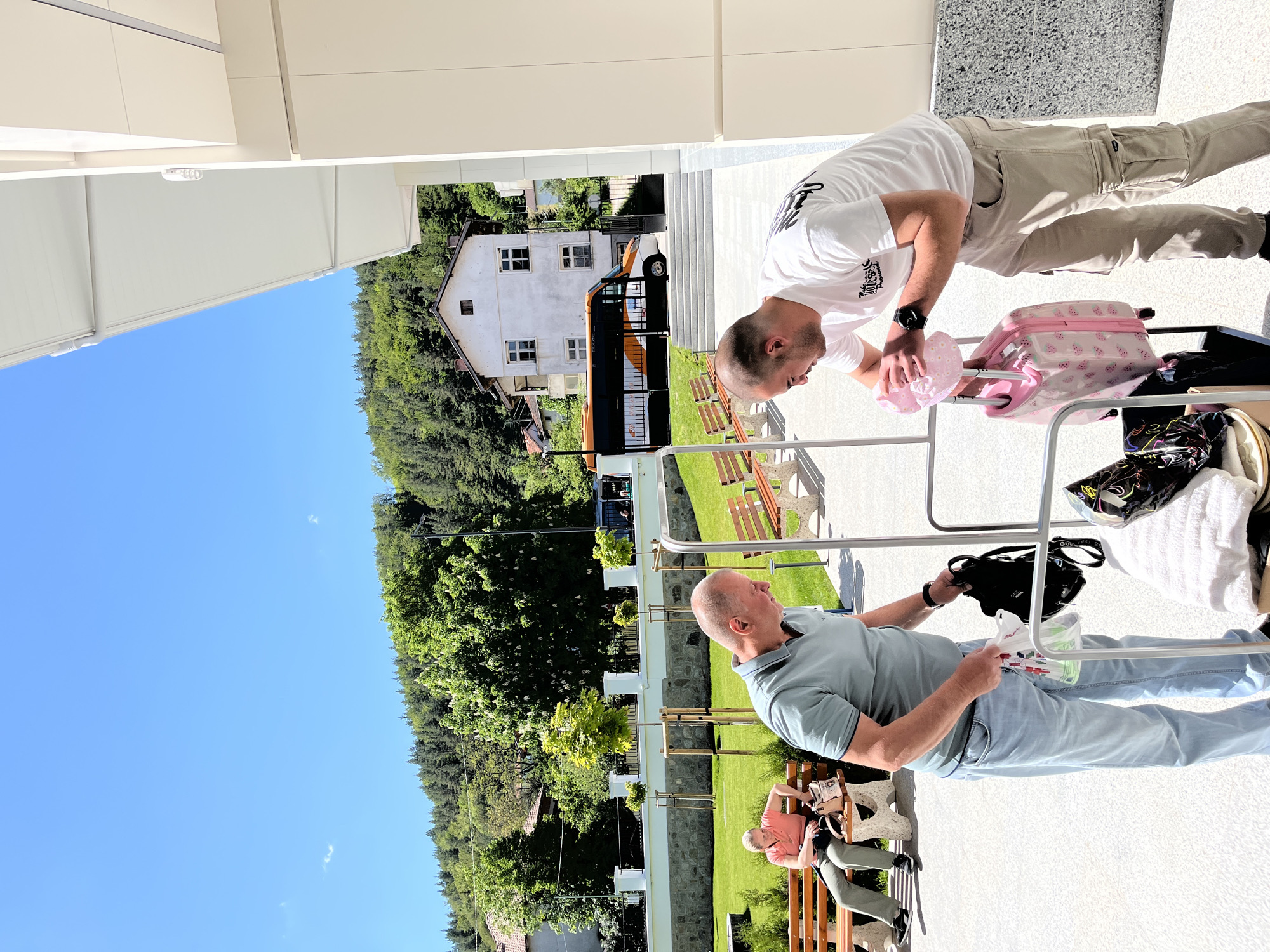 A vertically oriented image depicts two men in an outdoor setting, seemingly within the city of Belchinski Bani, Bulgaria, given the presence of houses and lush greenery characteristic of the region.
One man, clad in a light blue polo shirt and blue jeans, is seated on a bench. He holds a white object in his left hand and appears to be looking at it. A black object rests on the bench beside him. A second person is sitting on the same bench.
A second man stands near luggage, dressed in a white T-shirt and light brown cargo pants. He is bending over a pink suitcase, with what appears to be a pink hat in his hands.
The scene includes a house and a yellow and black bus in the background, set against a backdrop of trees and a clear blue sky. The environment includes a gray patterned surface, possibly concrete or stone, upon which the individuals and objects are situated. The scene has linear metal bars on the foreground. There are benches scattered around as well.