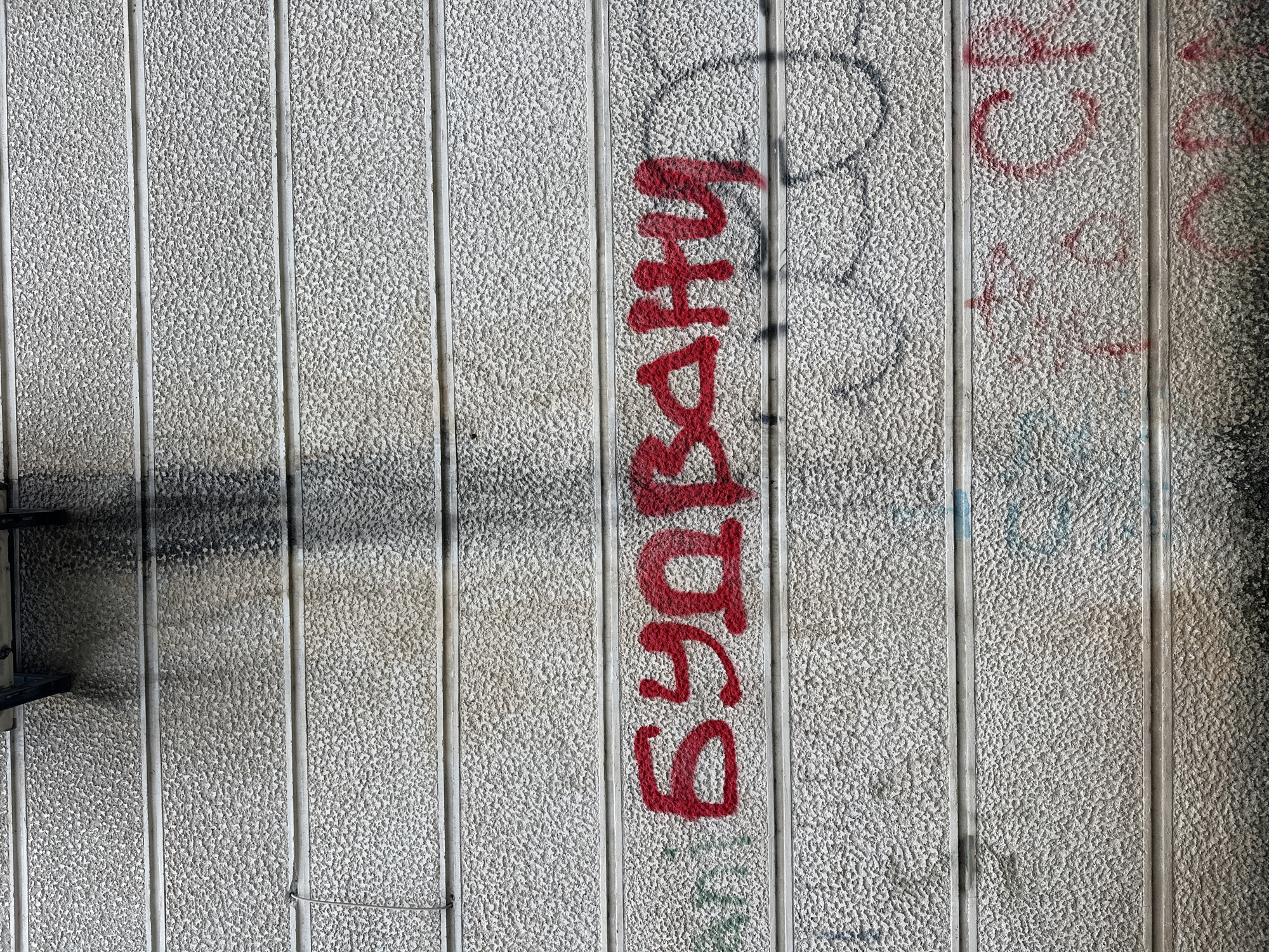 The image shows a close-up of a textured, concrete wall with vertical grooves. There is graffiti on the wall. The number "69" is painted in red, oriented vertically, followed by the word "BARRY" also in red and vertically oriented. Other graffiti, possibly in black and red, is also visible, but less clear. The wall is gray-white with a speckled texture. Some smudges and discoloration mark the wall, near the top. A piece of metallic hardware is partially visible on the left edge of the image. The location is Budva, Montenegro.