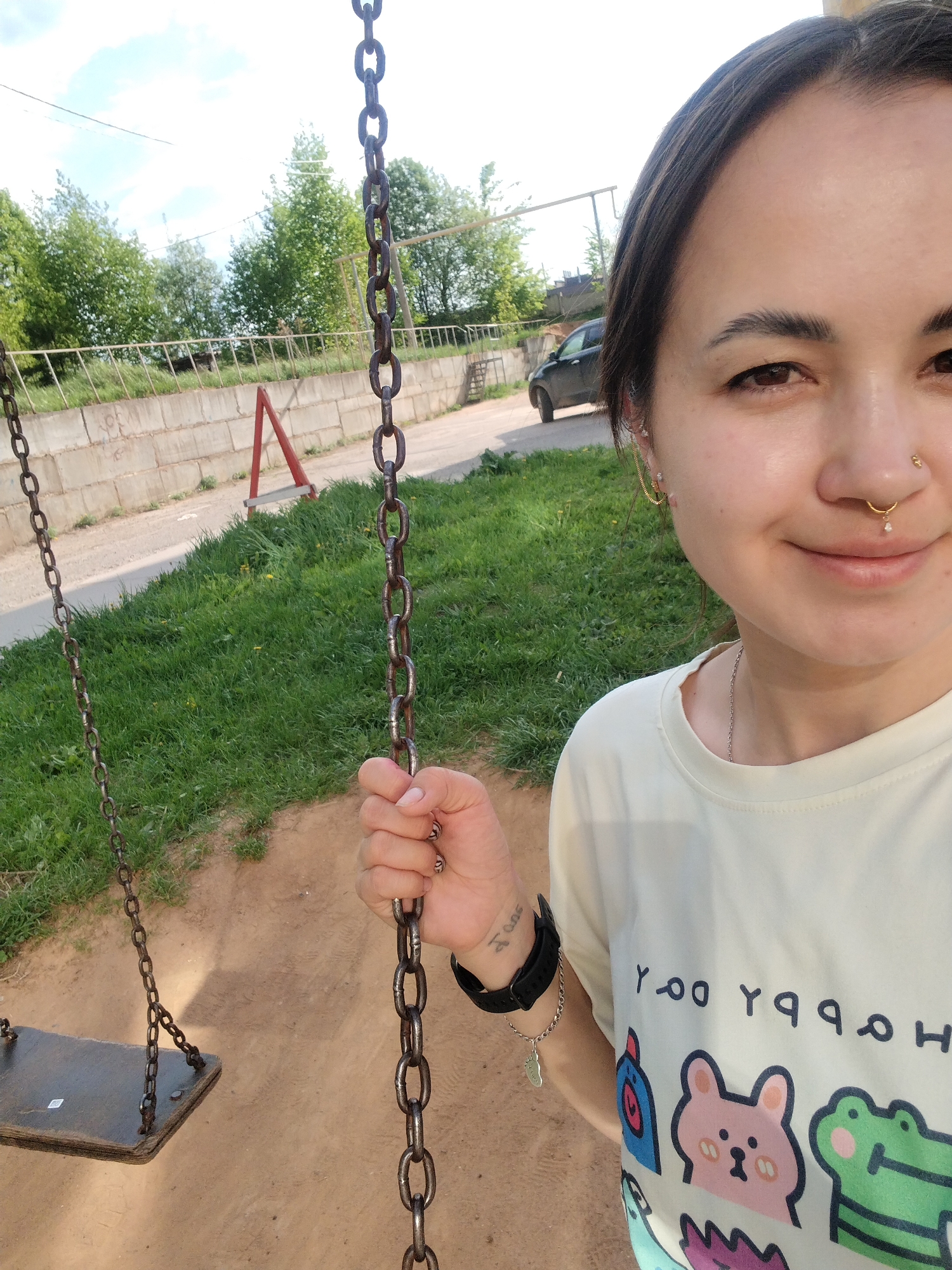 A selfie shows a young woman on a swing in Noviy Urengoy, Russia. She is holding the swing chain, smiling at the camera. She wears a white shirt with cartoon animal prints. A watch and a bracelet are visible on her left wrist. The background includes grass, a wall, a road, a car, and trees. A red safety barrier is visible in the distance.