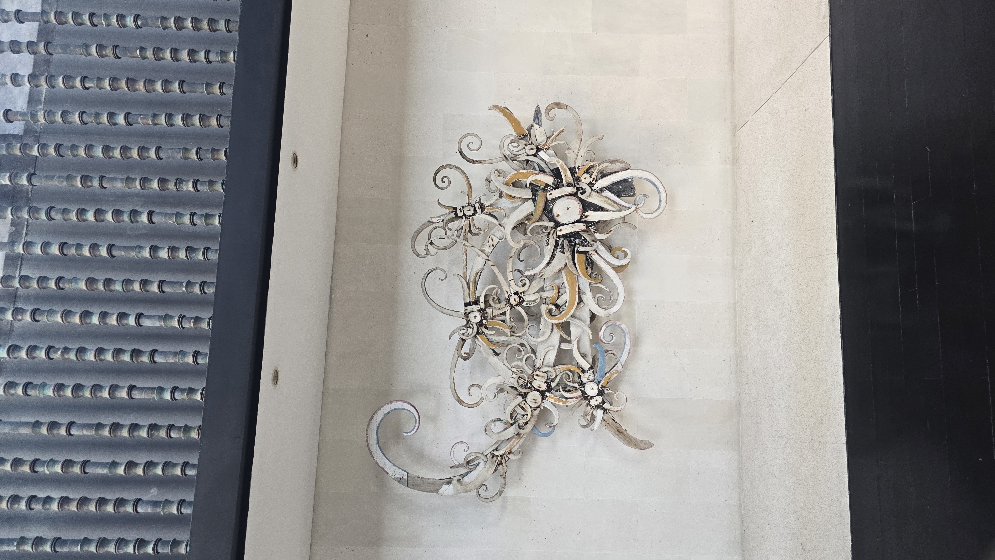 The image shows an ornate, metal floral wall decoration on a textured, light-colored wall, possibly made of tiles, next to a dark gray corrugated metal or wooden wall. The overall setting appears to be indoors. It is located in Kuta Selatan, Indonesia.