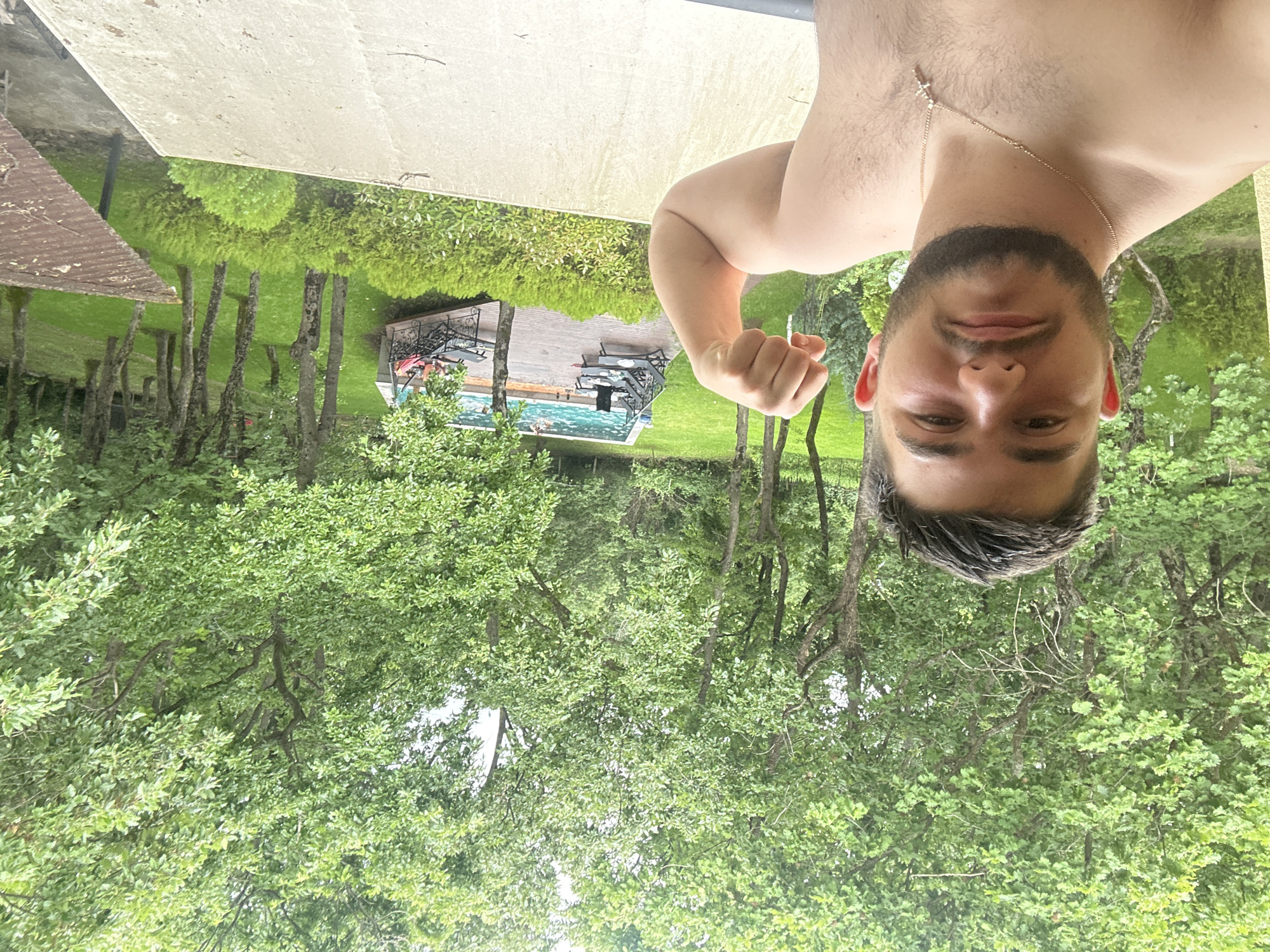 A bare-chested man is captured in a selfie-style shot outdoors, appearing to hold the camera from above. He is seen from the chest up, looking directly at the viewer with a neutral expression. He has short, dark hair and a beard, and wears a thin gold chain with a small cross pendant around his neck. The man is flexing his right arm, making a fist to show his bicep.
Behind him, a vibrant outdoor scene unfolds. In the mid-ground, a lush green lawn is dotted with numerous trees featuring slender trunks and abundant green foliage. Further back, a blue swimming pool is visible, bordered by a wooden deck furnished with outdoor tables and chairs. To the left of the pool area, a building with a reddish-brown tiled roof can be seen. The immediate foreground below the man suggests he is on an elevated position, possibly a balcony or terrace, indicated by a light-colored concrete surface.
The image appears to be taken during daytime, under bright, possibly overcast conditions, characteristic of a pleasant summer or spring day. The setting evokes a sense of relaxation, likely in a private garden or resort area. There is no visible text. The location context for this scene is Burgas, Bulgaria.