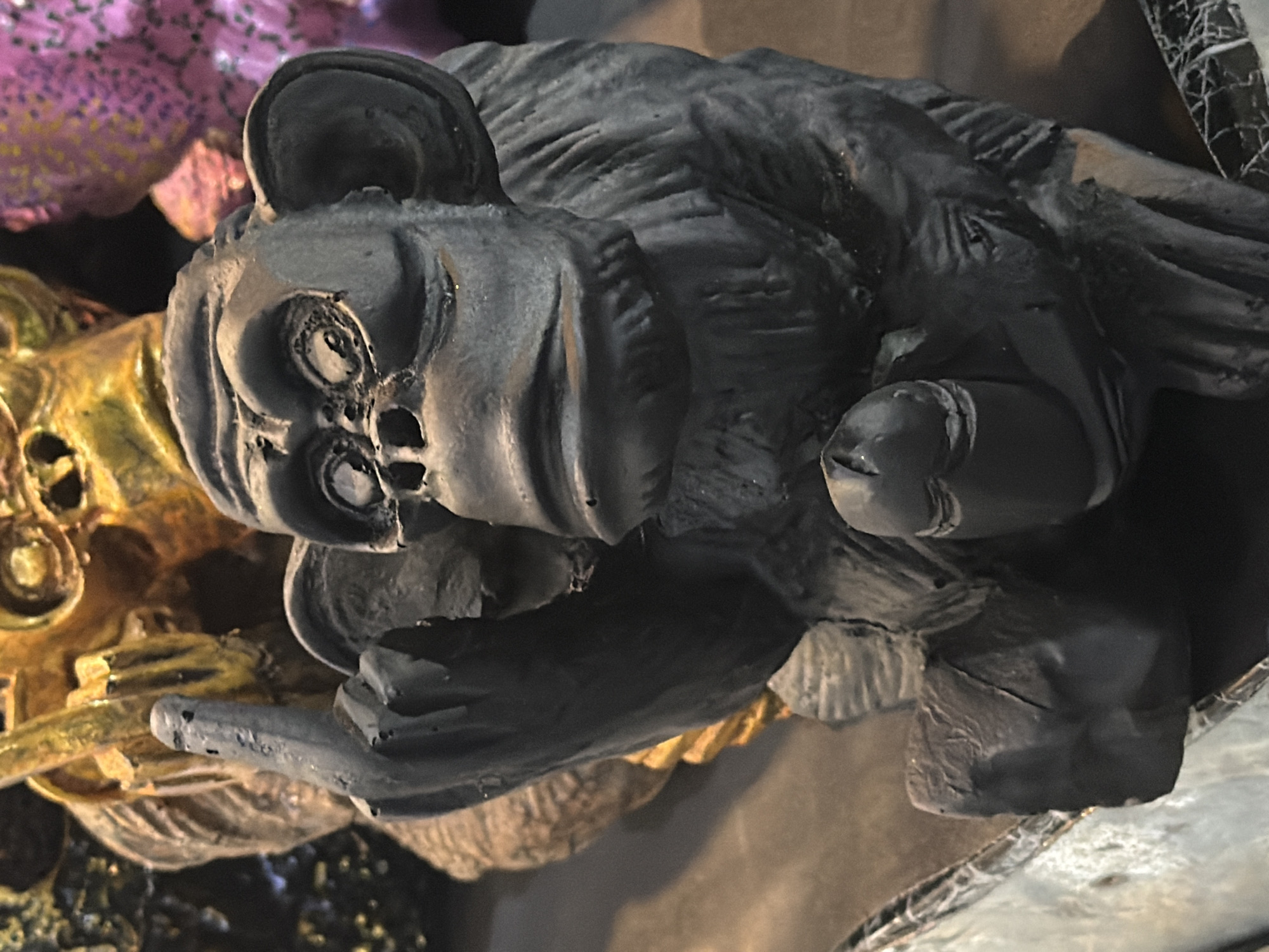 The image displays a close-up, high-angle shot of a dark, possibly Halloween-themed, figurine. The figurine appears to be made of a matte material, possibly plastic or resin, and is painted a dark, almost black color with some subtle reddish-brown undertones visible in the creases and textures.
The subject of the figurine is a stylized, somewhat monstrous creature with large, circular ears. Its face is prominent, featuring two large, hollow circular eyes with dark centers. The nose area is sculpted with indentations and ridges, and the mouth is a dark slit. The texture of the figurine suggests a rough, weathered, or perhaps even furry appearance on its body, with sculpted lines and furrows creating a sense of depth and texture. One of its arms is extended forward, with a hand or paw also sculpted with detail. The figurine is positioned at an angle, resting on or near other objects.
Behind the main figurine, glimpses of other objects can be seen. To the left, there is a brightly colored object with a textured surface, appearing to be a mix of purple and pink hues, possibly another decorative item or part of a larger display. Below and to the left of the main figurine, a golden-colored object with intricate sculpted details is visible, adding a contrasting color and texture to the scene. The background is dark and indistinct, suggesting an indoor setting or a dimly lit environment.
No people are visible in the frame. The activity appears to be a static display of decorative figurines. Notable details include the rough, textured finish of the main figurine and the contrasting colors of the surrounding objects. There is no visible text in the image. The overall impression is one of a Halloween or gothic-themed decoration. The location context of Kuta, Indonesia, does not provide any immediately apparent visual cues within the image itself.